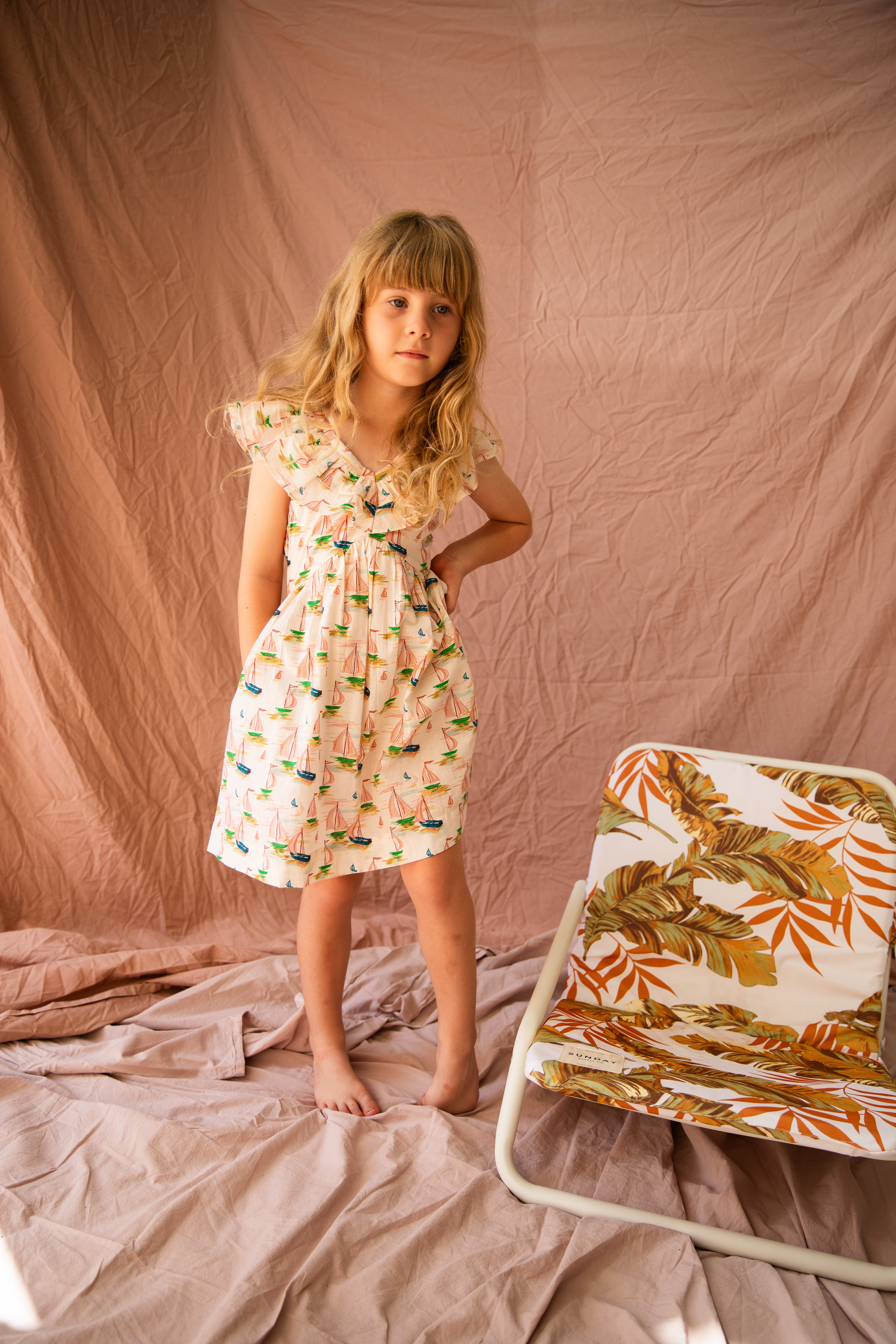 A young girl with long blonde hair stands barefoot on a crumpled pink fabric background, showcasing a breezy summer look in the Seychelles Dress Regatta by BELLA + LACE. With one hand on her hip, she exudes confidence next to a small chair draped in a regatta print that perfectly complements her outfit.