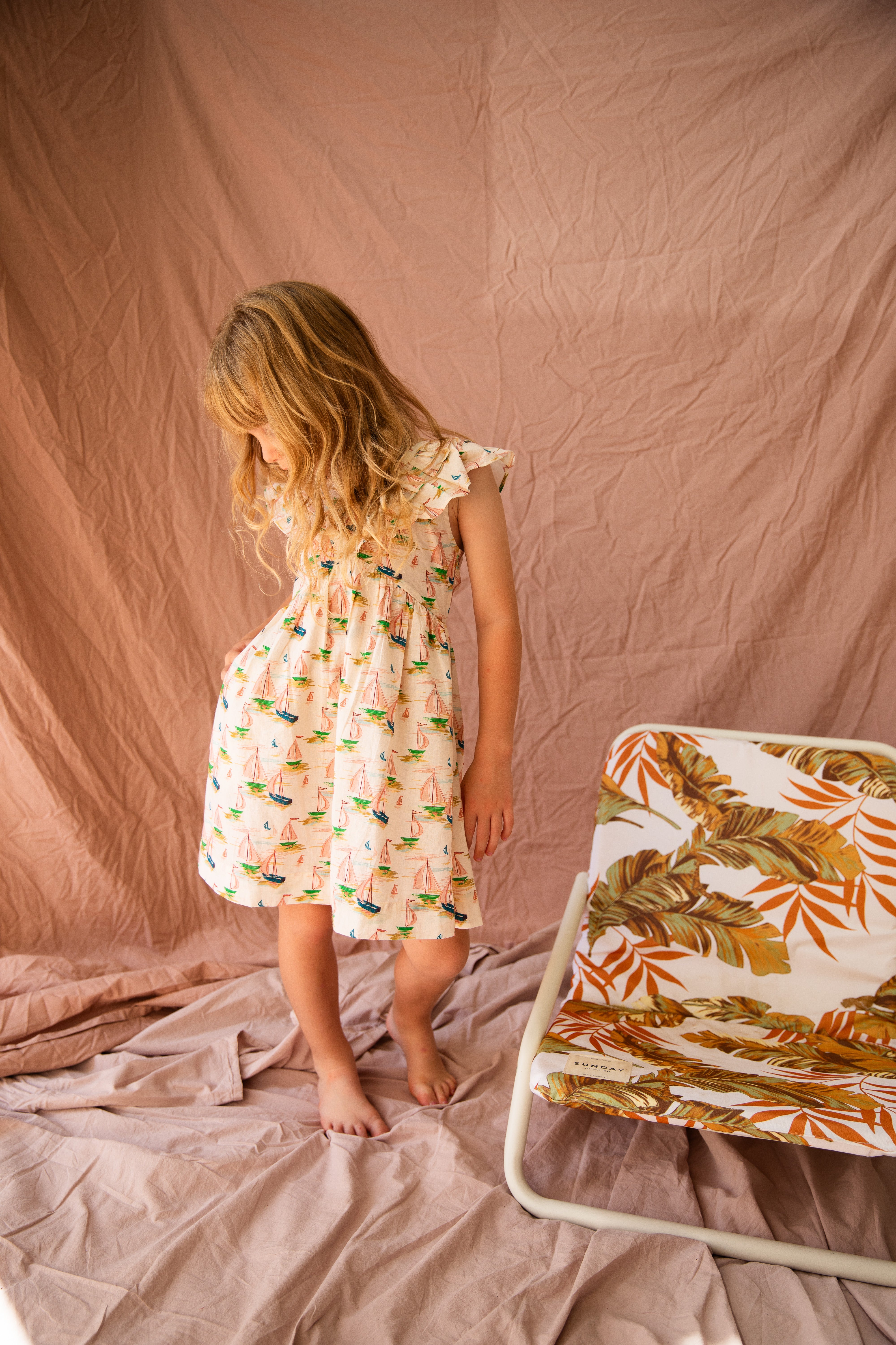 A young girl with long blonde hair wears the Seychelles Dress Regatta by BELLA + LACE, a perfect summer outfit crafted from light cotton voile. She stands barefoot on a soft fabric backdrop, next to a chair adorned with tropical leaf and regatta print fabric, enhancing the warm and relaxed setting.