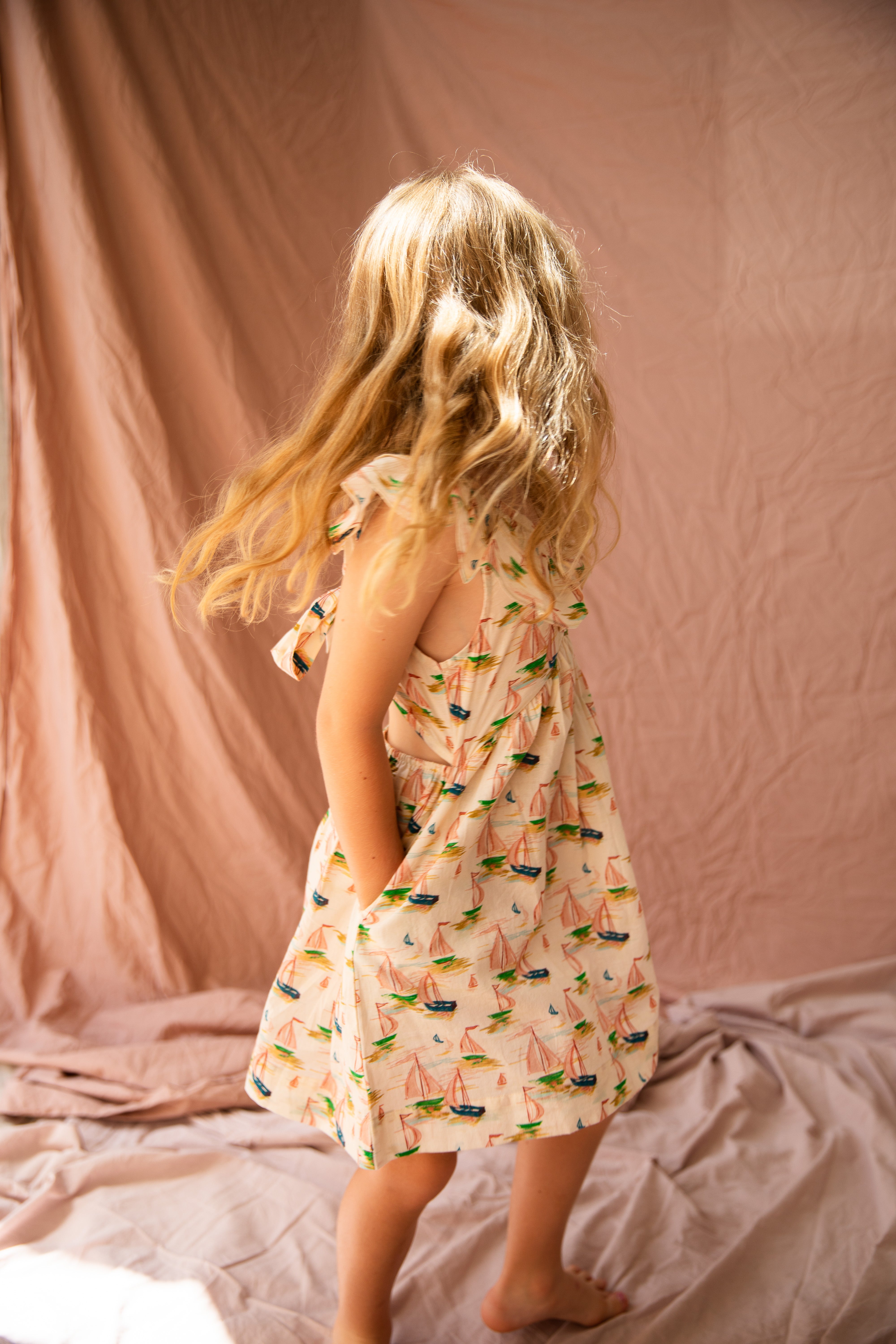 A young girl with long blonde hair stands barefoot on a soft fabric in a brightly lit room, wearing the sleeveless Seychelles Dress Regatta from BELLA + LACE, featuring a vibrant regatta print ideal for a summer outfit. The background consists of crumpled light pink drapery.
