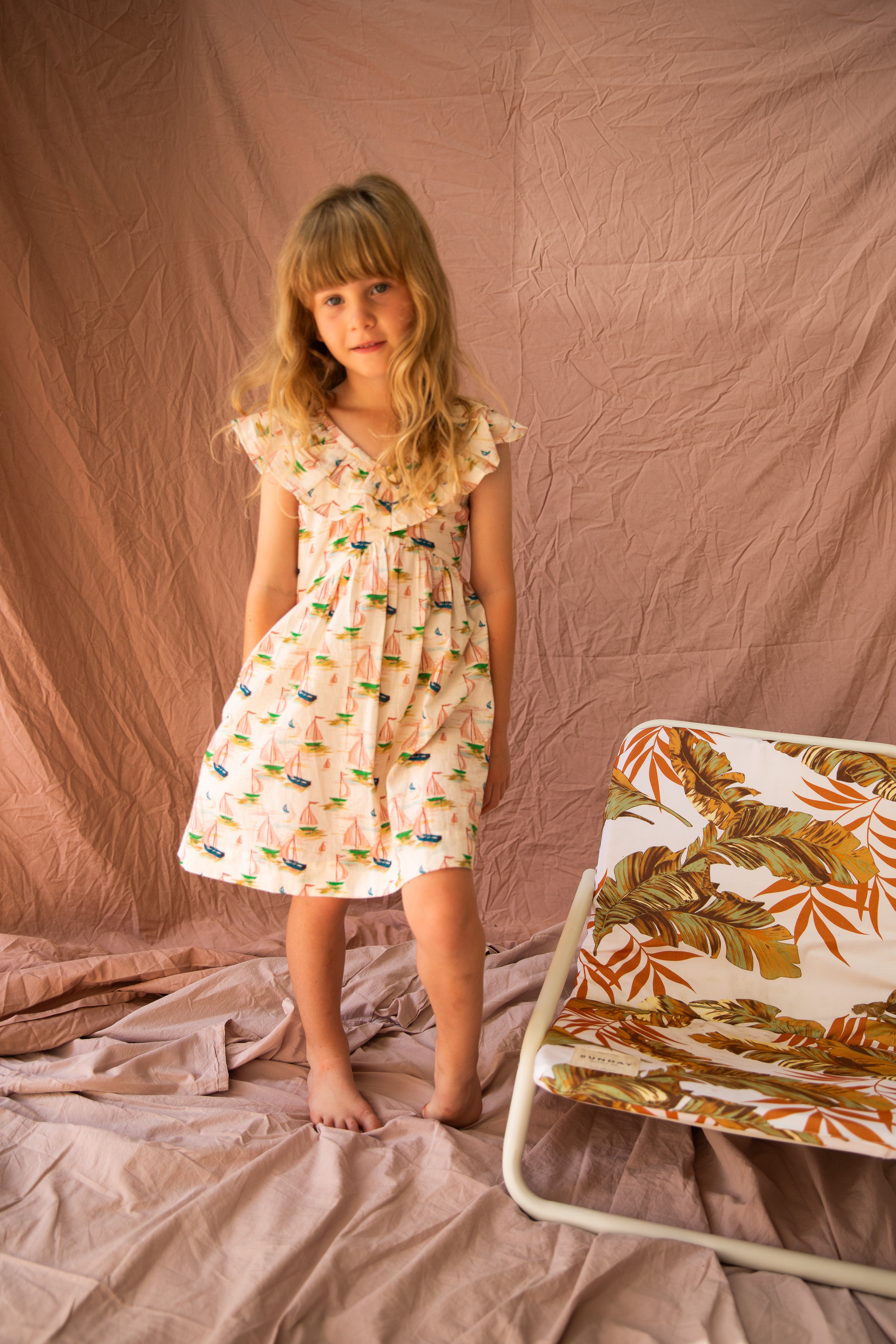 A young girl with long blonde hair stands barefoot on a fabric-draped floor, wearing the Seychelles Dress Regatta by BELLA + LACE—a summer outfit crafted from airy cotton voile featuring a charming regatta print. Beside her, a small chair showcases a tropical leaf design against soft, earthy-toned curtains.