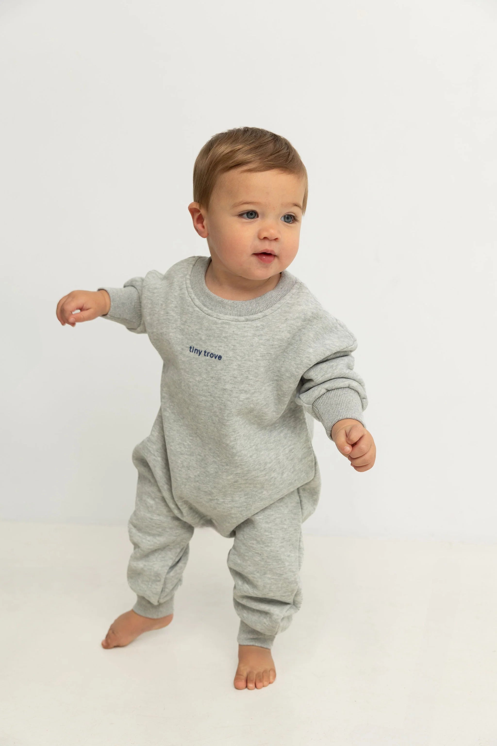 A toddler with light brown hair stands barefoot on a white floor wearing the TINY TROVE Tiny Trove - Otto Puff Circle Logo Romper in Grey Marle, against a plain white background.
