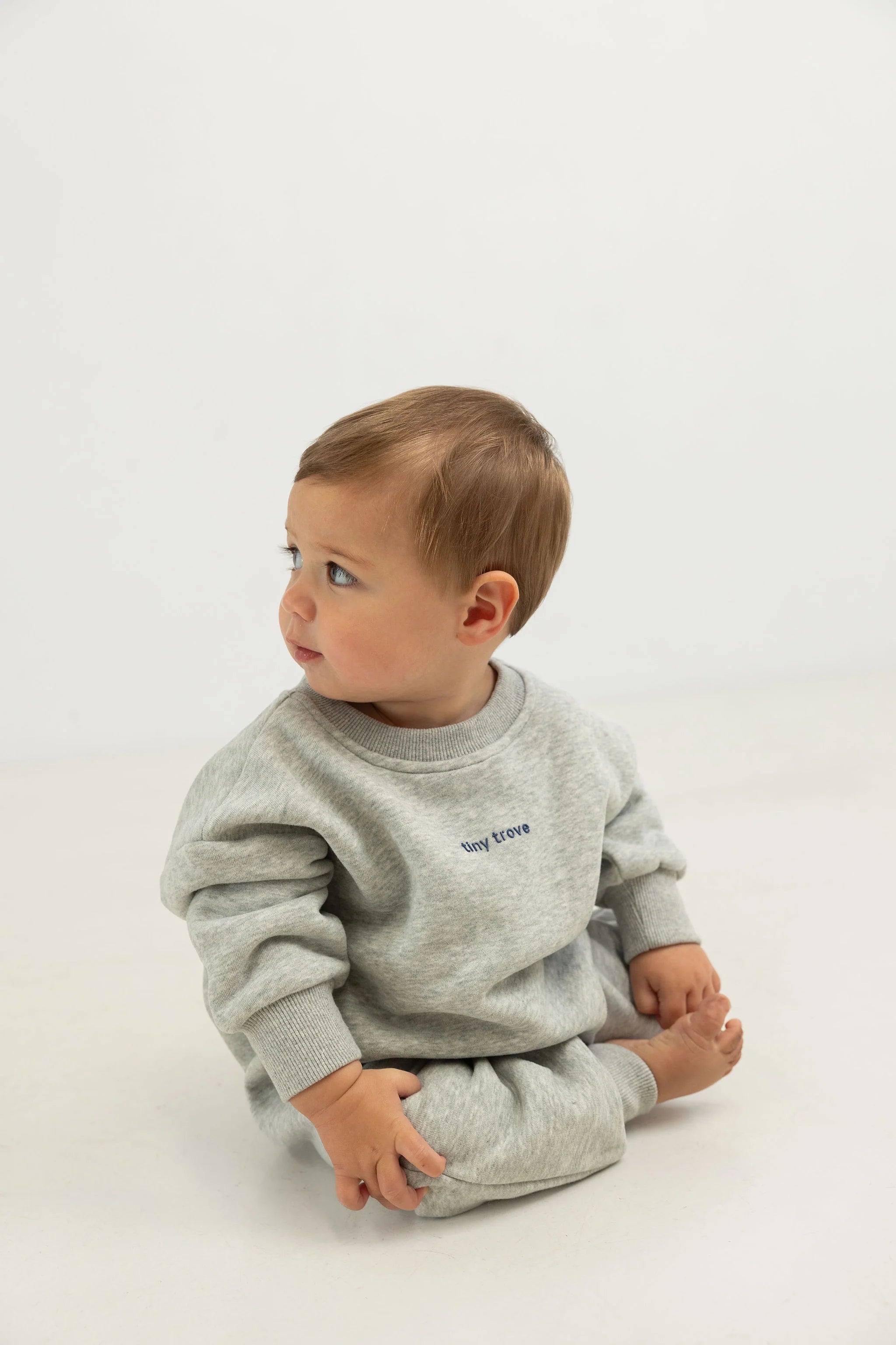 A young child with light brown hair sits barefoot on a light surface, wearing the TINY TROVE Otto Puff Circle Logo Romper in Grey Marle, featuring 3D logo detailing. The background is plain white.
