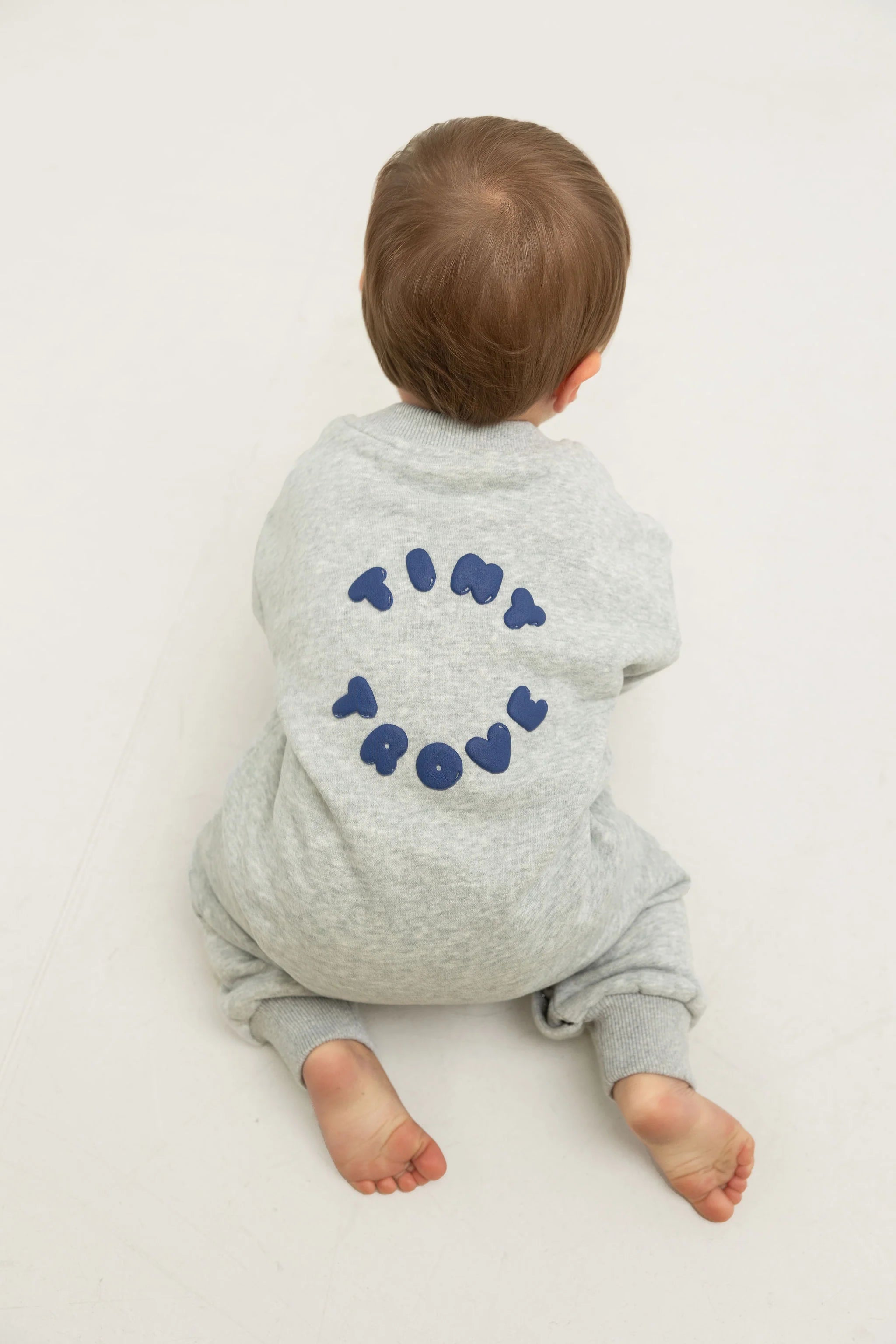 A young child sits barefoot on a pale floor, wearing the TINY TROVE Otto Puff Circle Logo Romper in Grey Marle, featuring subtle 3D logo detailing and “TINY TROUBLE” text on soft cotton fleece.