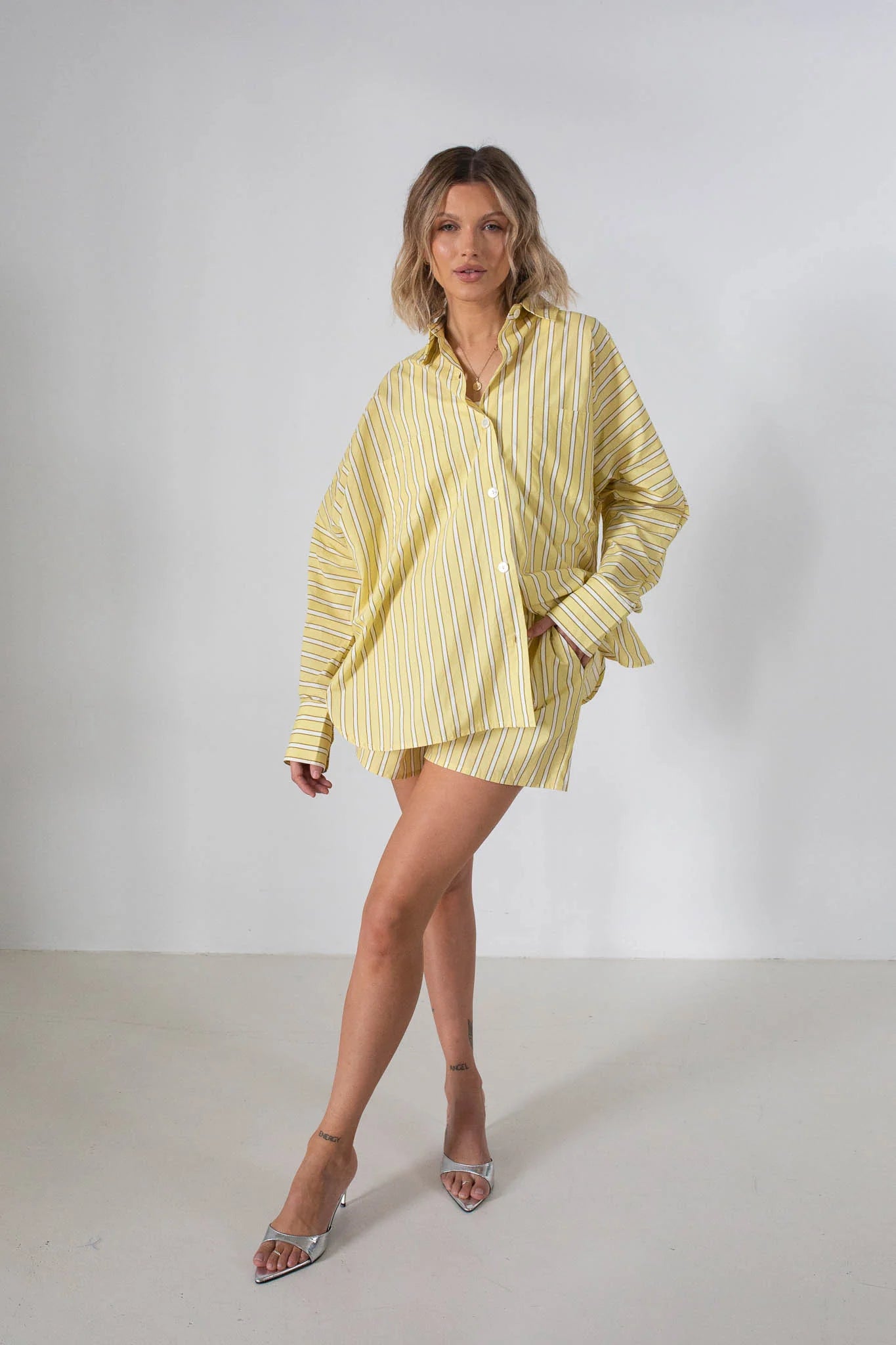 A woman poses indoors against a white background wearing the TINY TROVE Tiny Trove Women's Isla Stripe Cotton Set in yellow and white stripes, paired with clear high-heeled shoes. Her blonde hair is styled in waves for a relaxed, casual look.