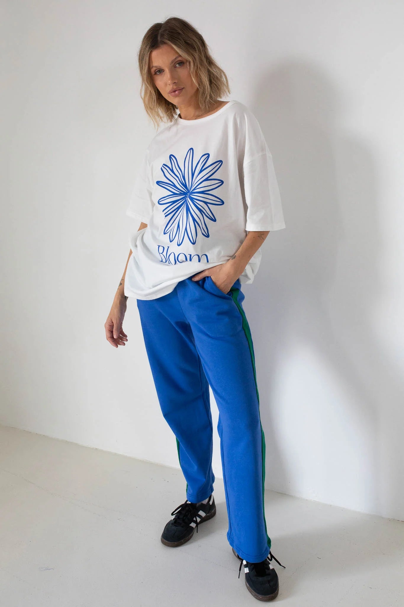 A woman wears the TINY TROVE Tiny Trove - Women's Bloom Relaxed Tee White/Blue, featuring an oversized organic cotton fit with a blue graphic flower and "bloom" logo, paired with blue track pants and black sneakers.