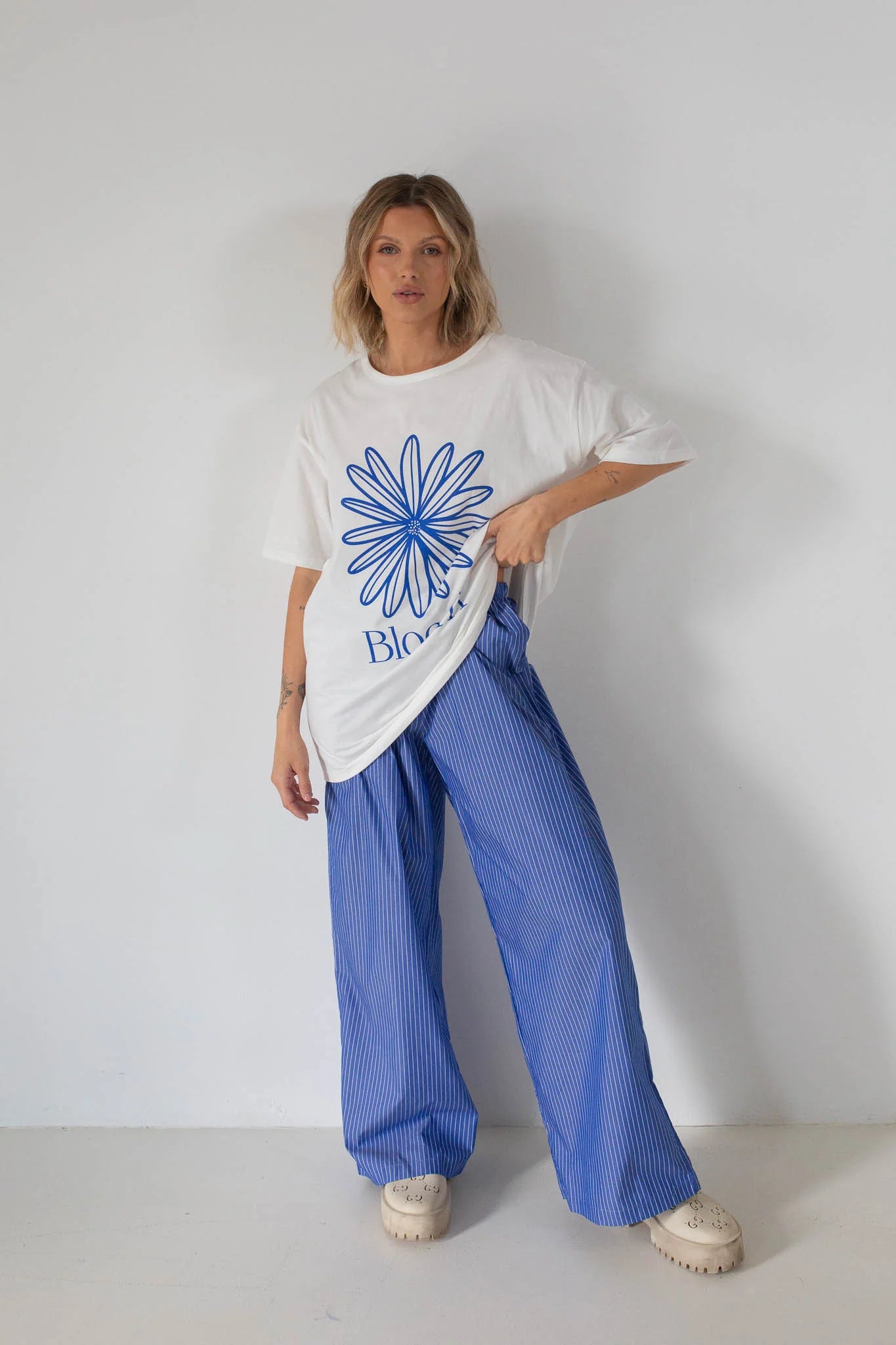 A person wears the Tiny Trove - Women's Bloom Relaxed Tee White/Blue by TINY TROVE, featuring a blue flower graphic and "Bloom" text, paired with wide-legged blue striped pants and beige chunky shoes against a plain white wall.