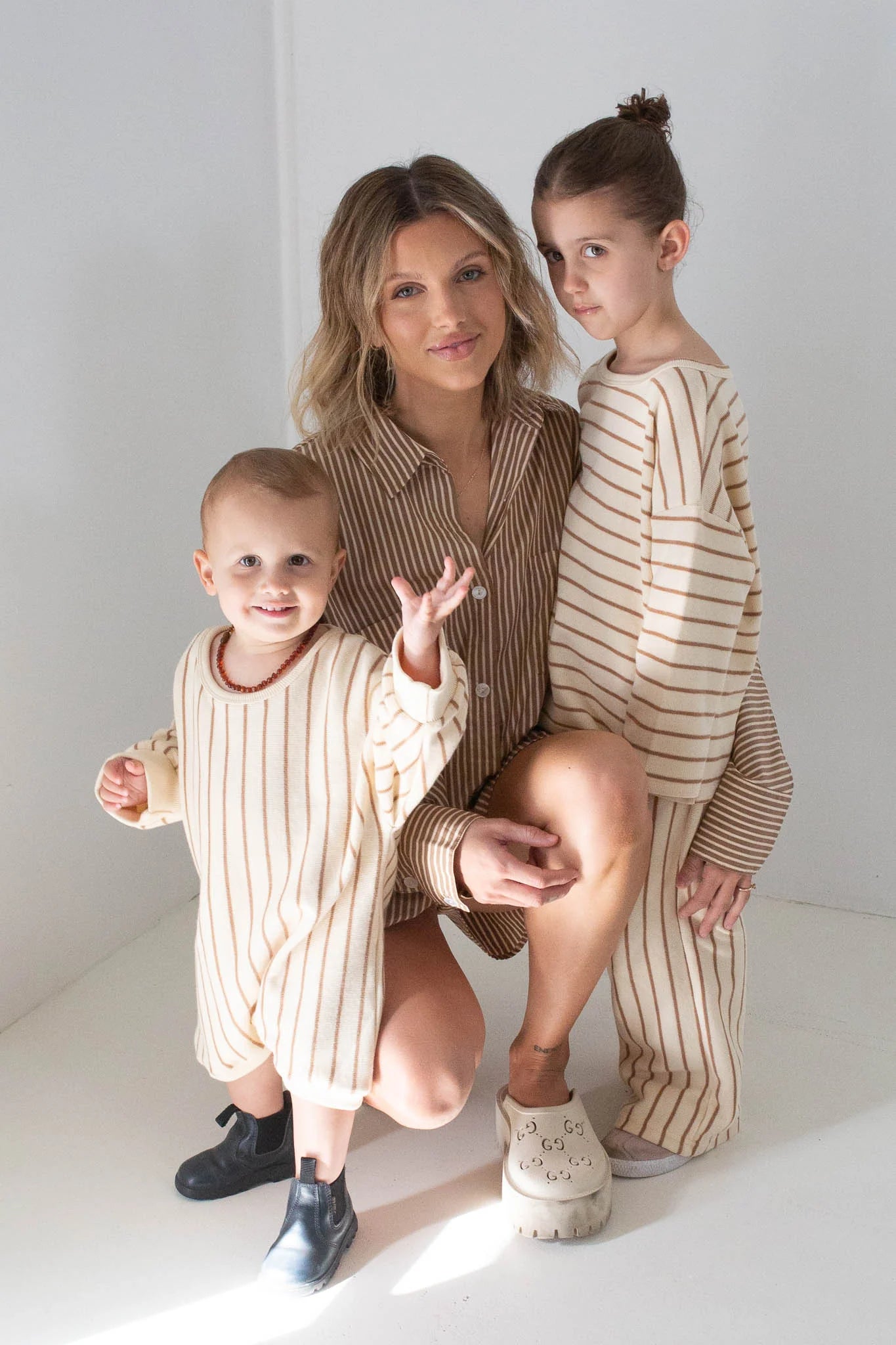 A woman kneels beside two young children in a sunlit room. All wear TINY TROVE’s Addie Cotton Stripe Romper in Cream/Latte Stripe, made from 100% cotton. The older child stands with hair in a bun; the younger smiles and waves in the cozy romper.