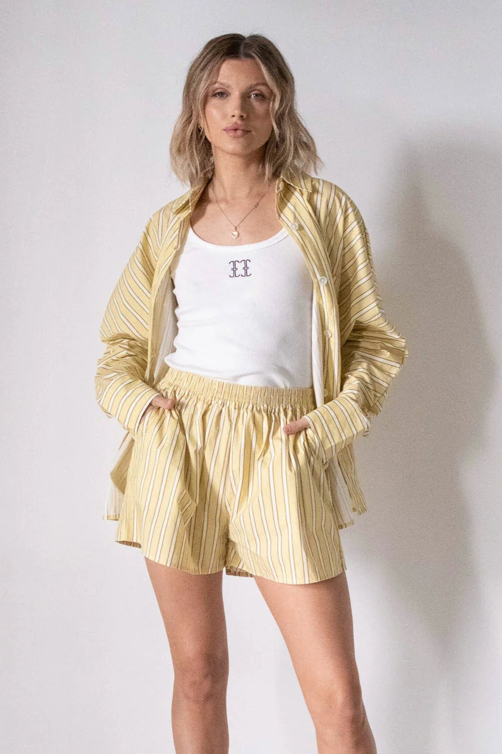 A woman with blonde, wavy hair poses against a plain background wearing the TINY TROVE - Women's Isla Stripe Cotton Set Yellow/White Stripe: a relaxed fit button-up shirt over a white tank and matching shorts.