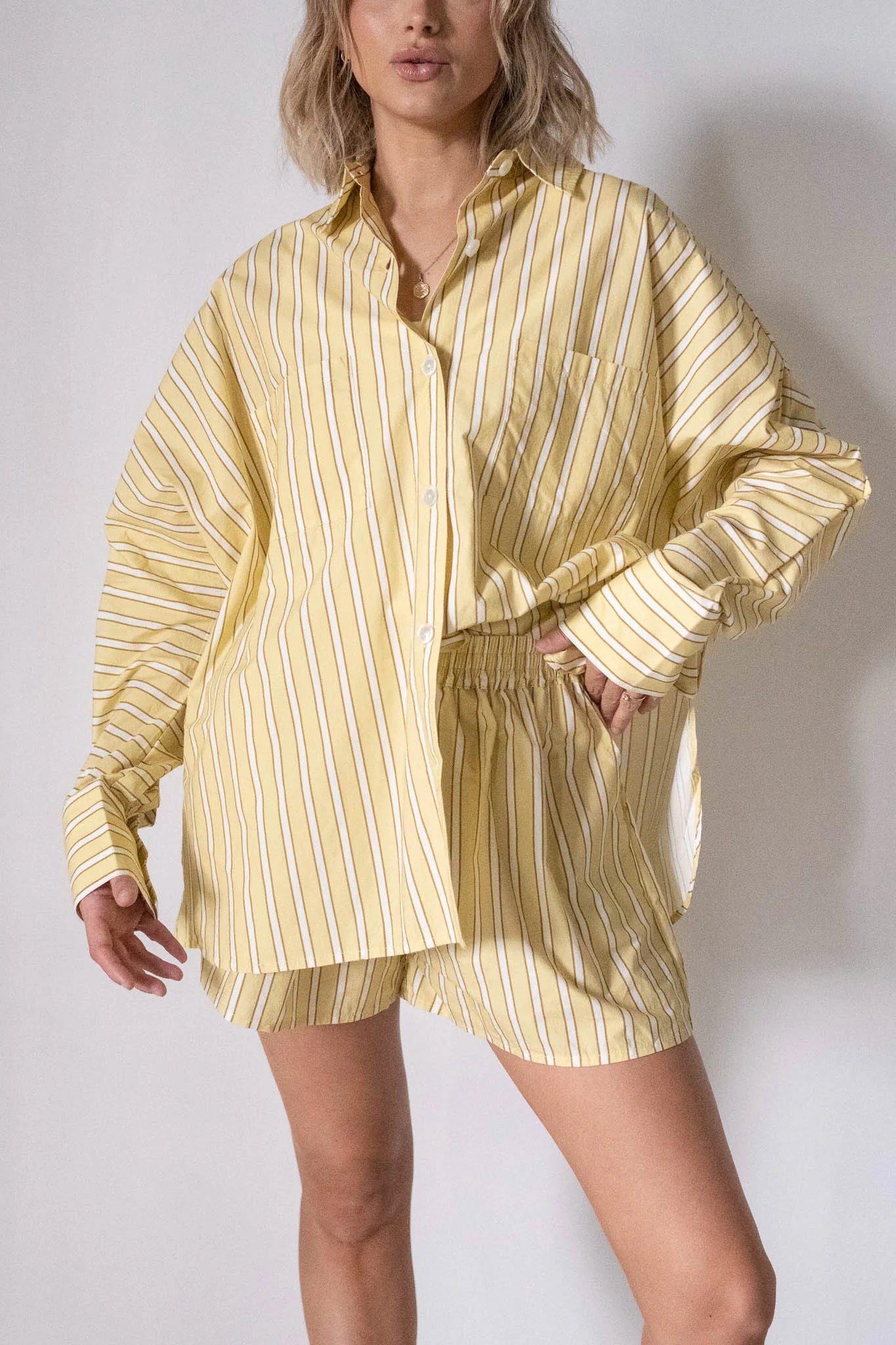 A person wears the TINY TROVE Women's Isla Stripe Cotton Set Yellow/White Stripe, a relaxed cotton matching set with a loose yellow button-up shirt and coordinating shorts for a casual vibe.