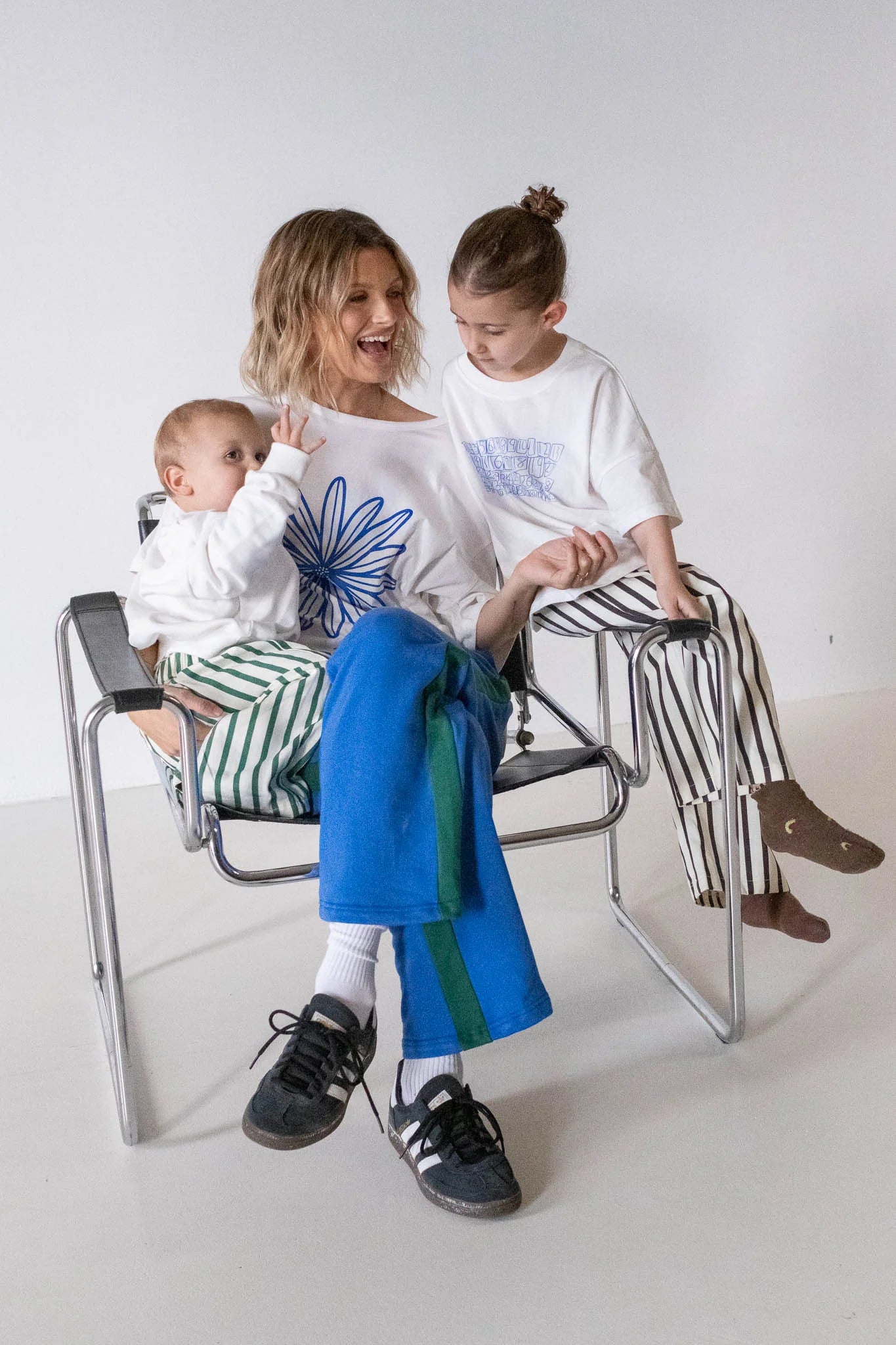 A smiling woman sits on a chair with her children, all wearing oversized, casual outfits adorned with stripes and playful logo prints. She wears the TINY TROVE Women's Bloom Relaxed Tee in White/Blue against a minimal white backdrop.