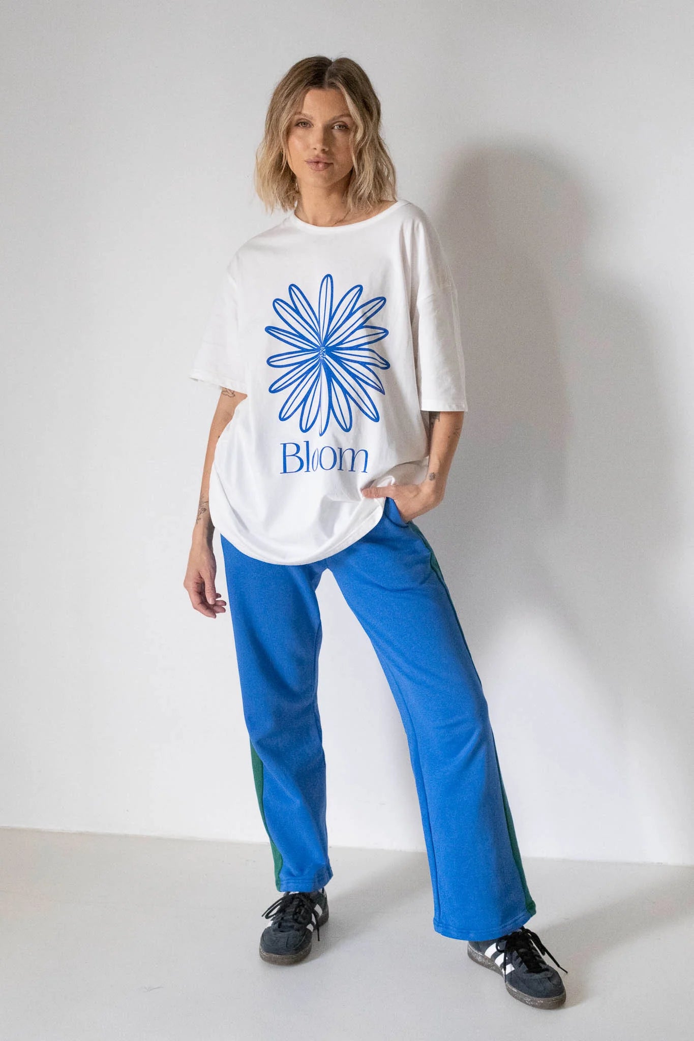 A person stands against a plain white wall, wearing the TINY TROVE Women's Bloom Relaxed Tee White/Blue with a blue geometric flower logo and "Bloom" print, paired with blue pants featuring green side stripes and black sneakers.