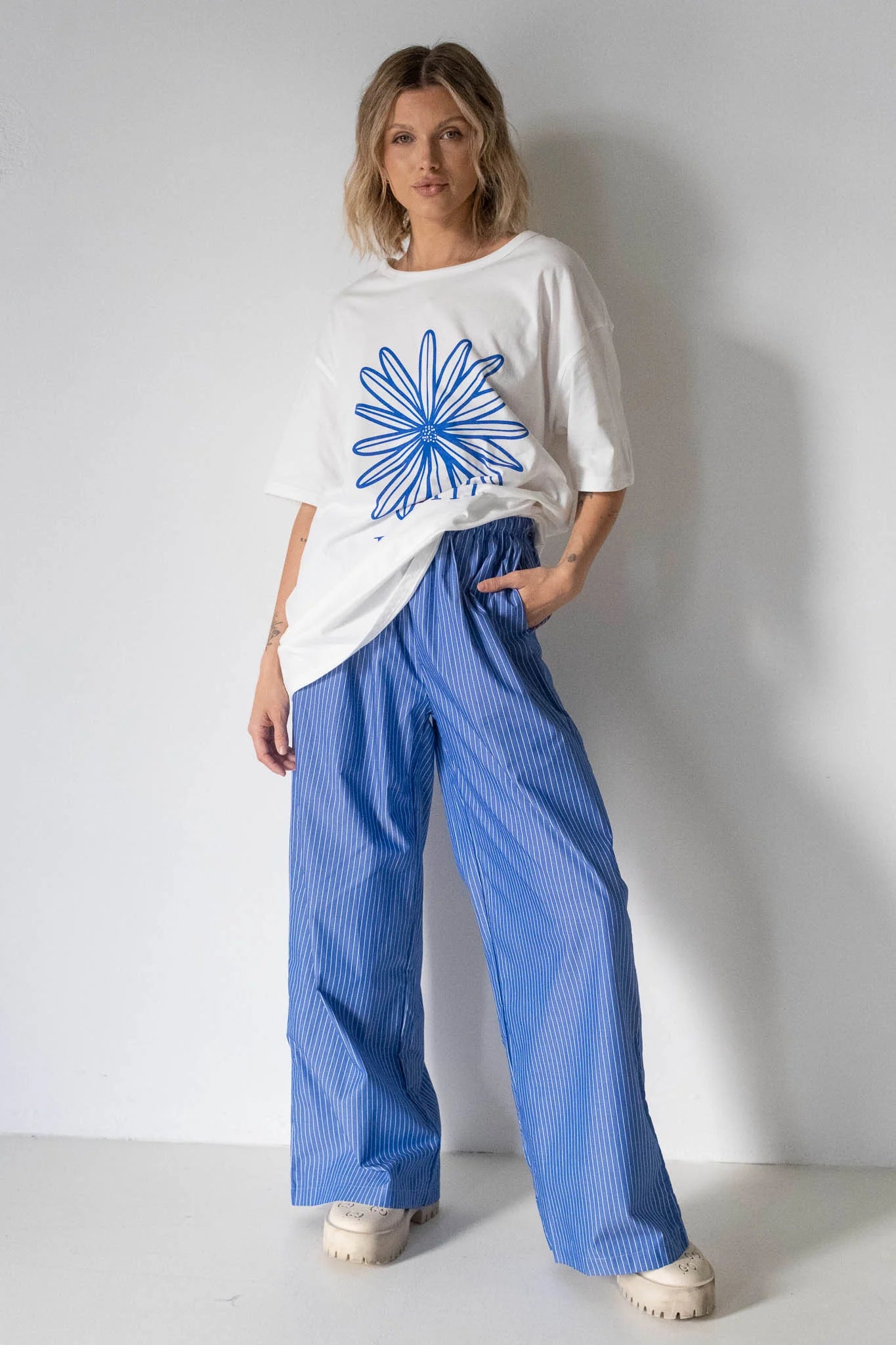 A person stands against a white wall wearing the TINY TROVE Tiny Trove - Women's Bloom Relaxed Tee White/Blue, featuring an oversized fit and large blue abstract flower graphic.