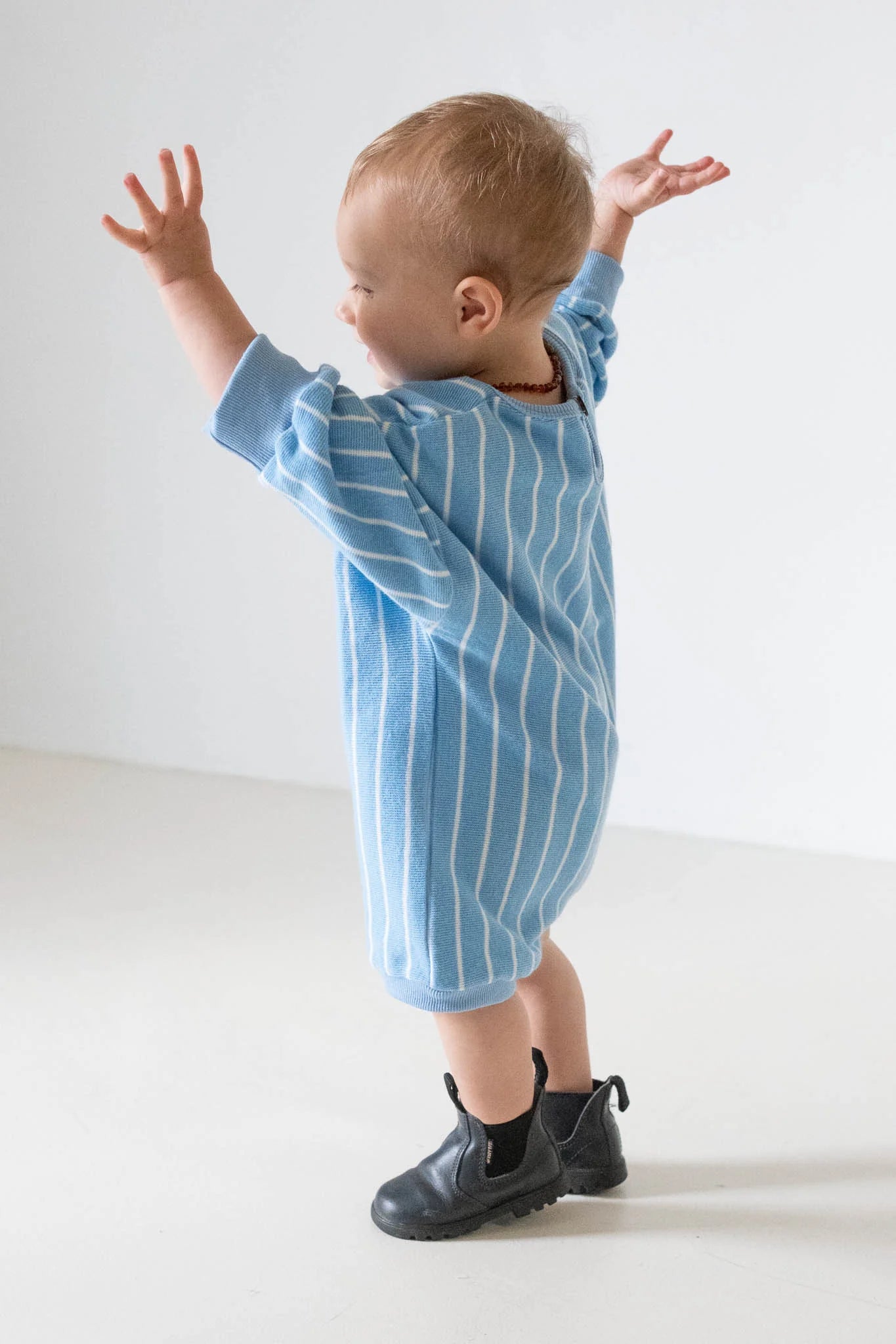 A toddler wearing the Tiny Trove - Addie Cotton Stripe Romper in blue/white stripe by TINY TROVE and black boots stands with arms raised, facing away from the camera against a plain white background.