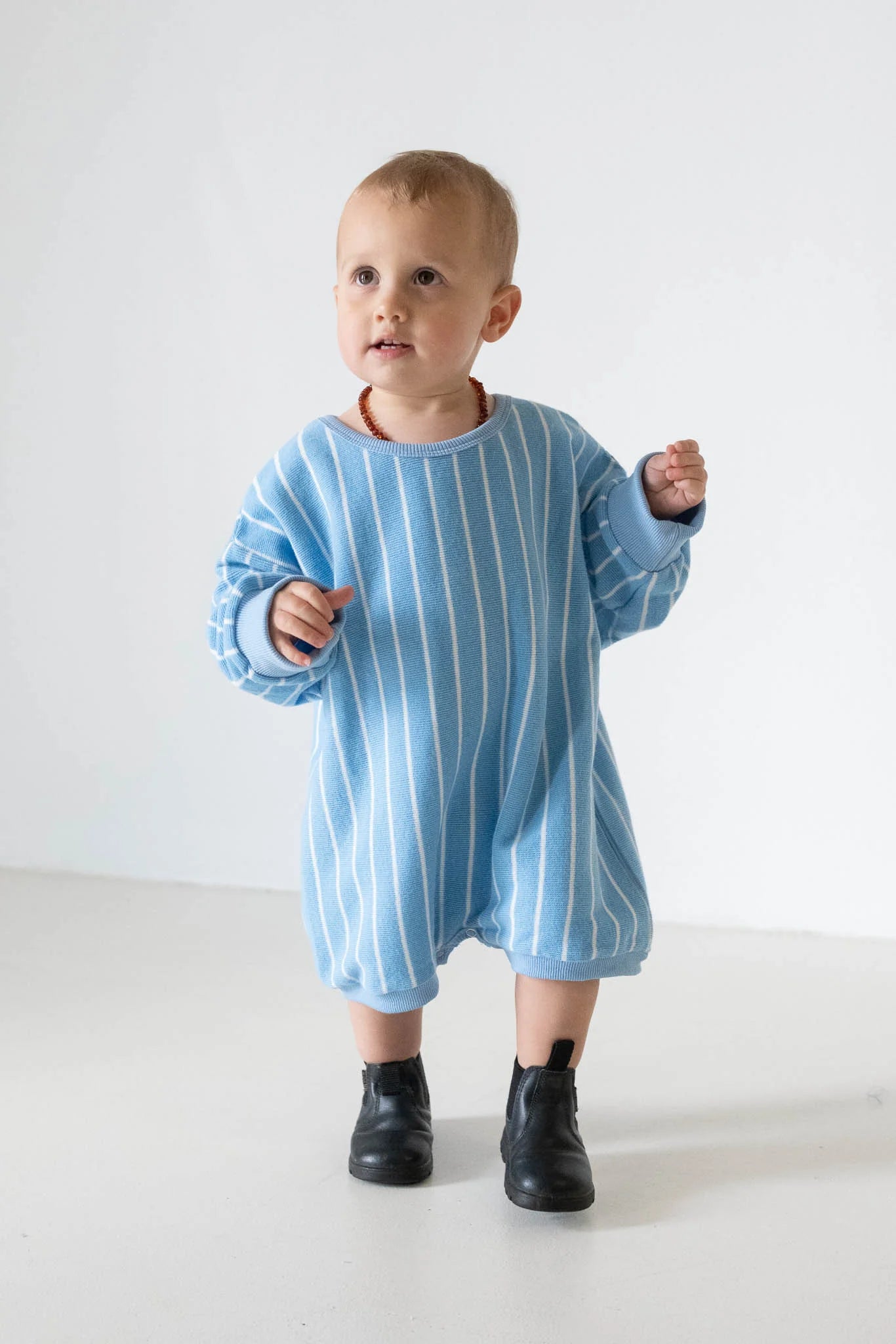 A toddler in the TINY TROVE Tiny Trove - Addie Cotton Stripe Romper Blue/White Stripe and black boots stands on a white floor against a plain white background.