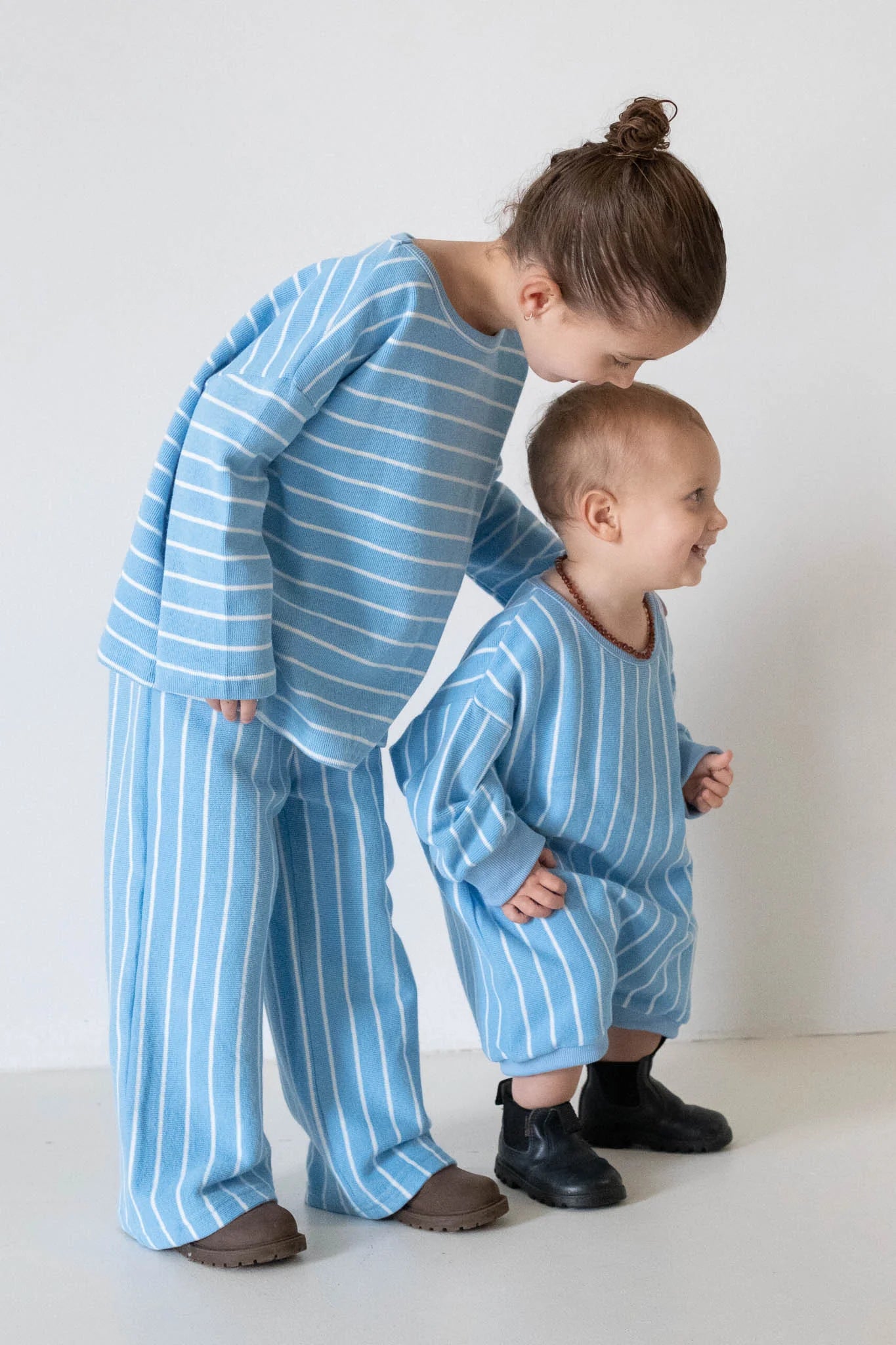 Two young children in matching blue and white outfits stand together. The younger smiles in a cozy TINY TROVE Addie Cotton Stripe Romper Blue/White Stripe, with shoes that look a little too big.