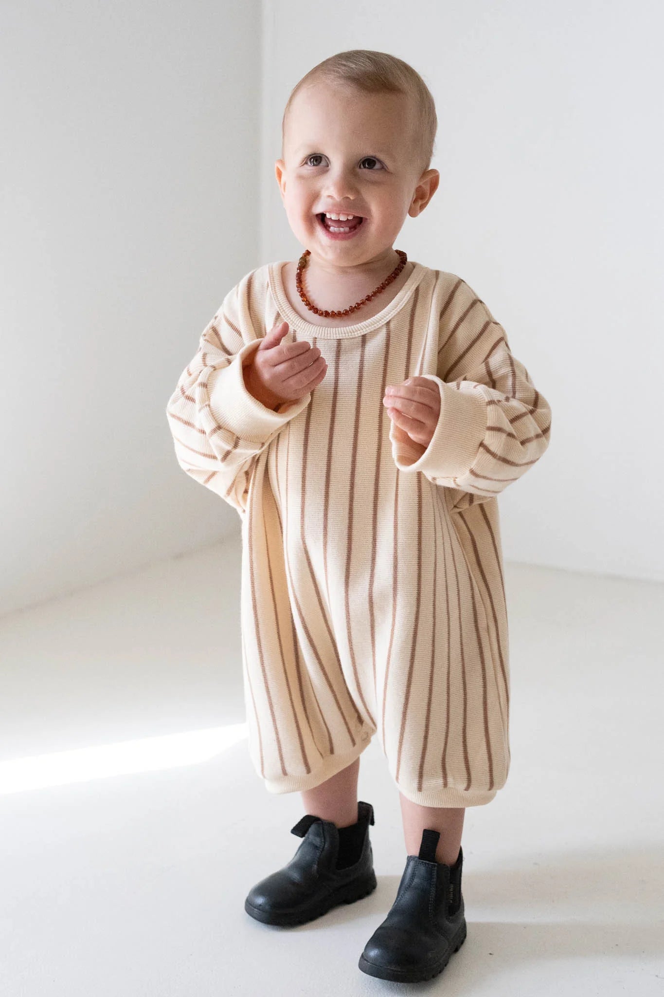 A smiling toddler wears the Tiny Trove - Addie Cotton Stripe Romper in Cream/Latte Stripe by TINY TROVE, standing in a bright, minimalist room.