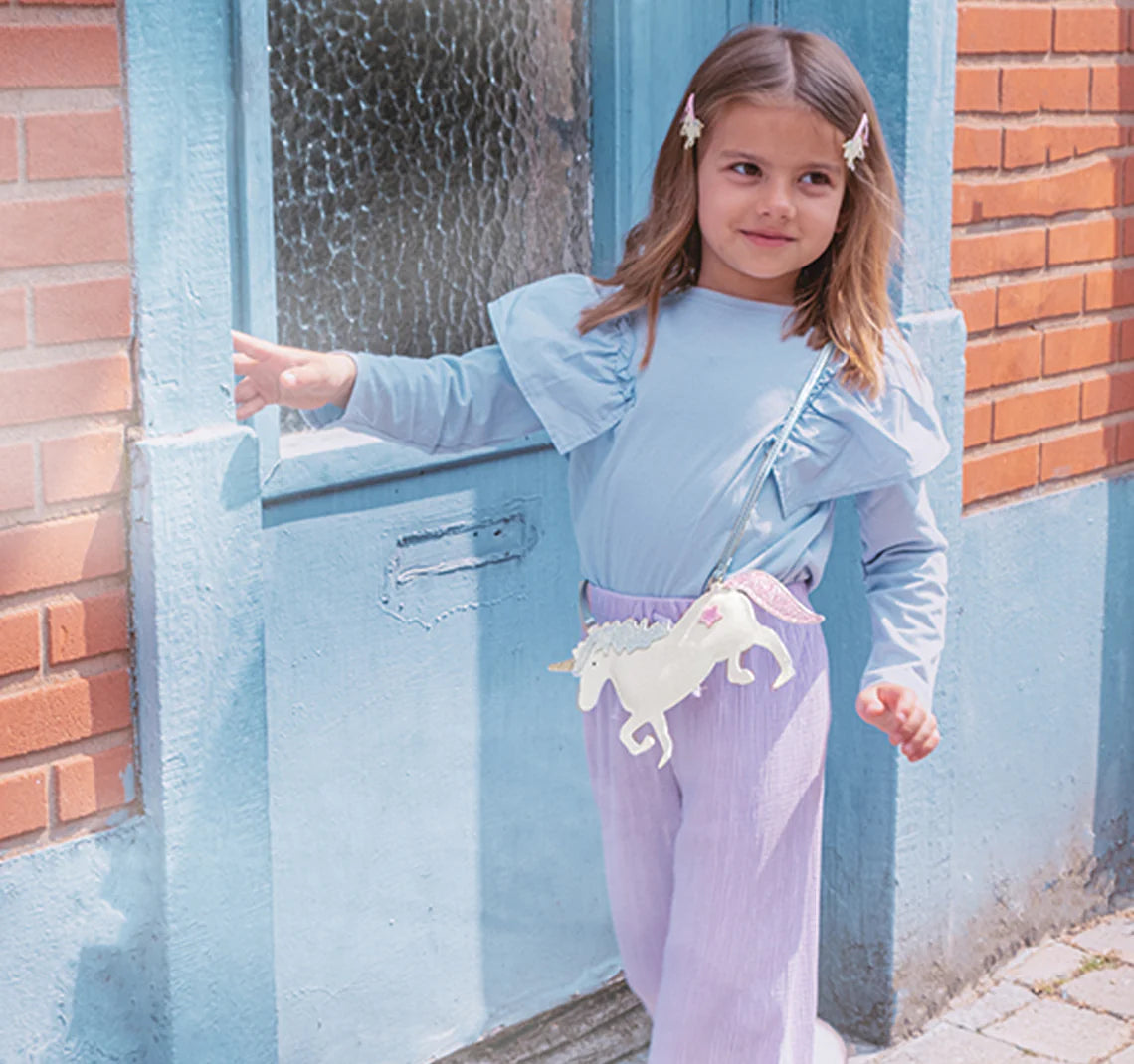 A young girl in a light blue ruffled top and lavender pants stands by a blue door, her hair clipped back and the ROCKAHULA Rockahula - Unicorn Glitter Bag with a metallic blue strap slung across her body.