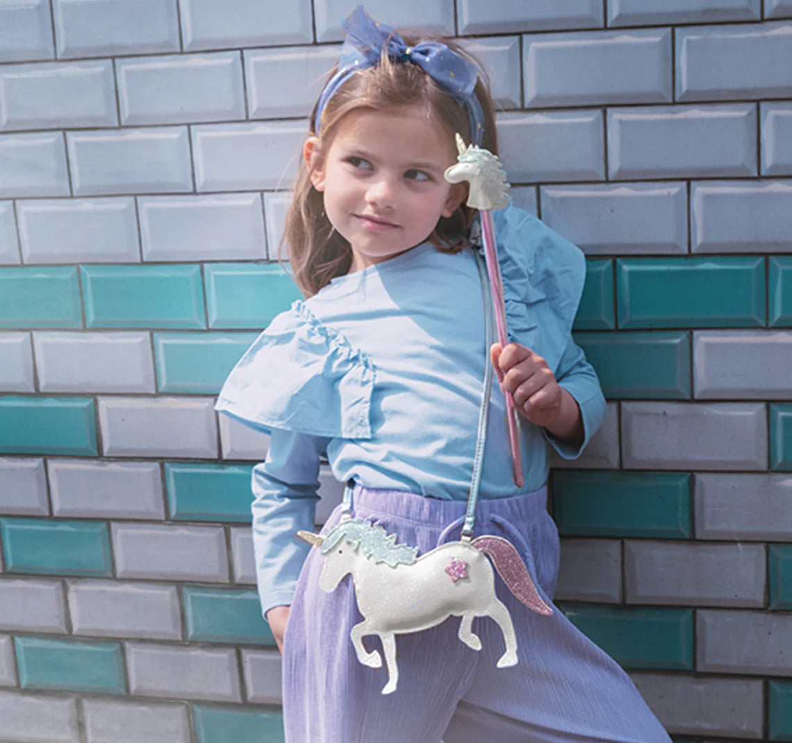 A young girl smiles and looks to the side, wearing the ROCKAHULA Rockahula - Unicorn Glitter Bag with a metallic blue strap, paired with a light blue blouse and headband, standing against teal and gray tiled bricks.