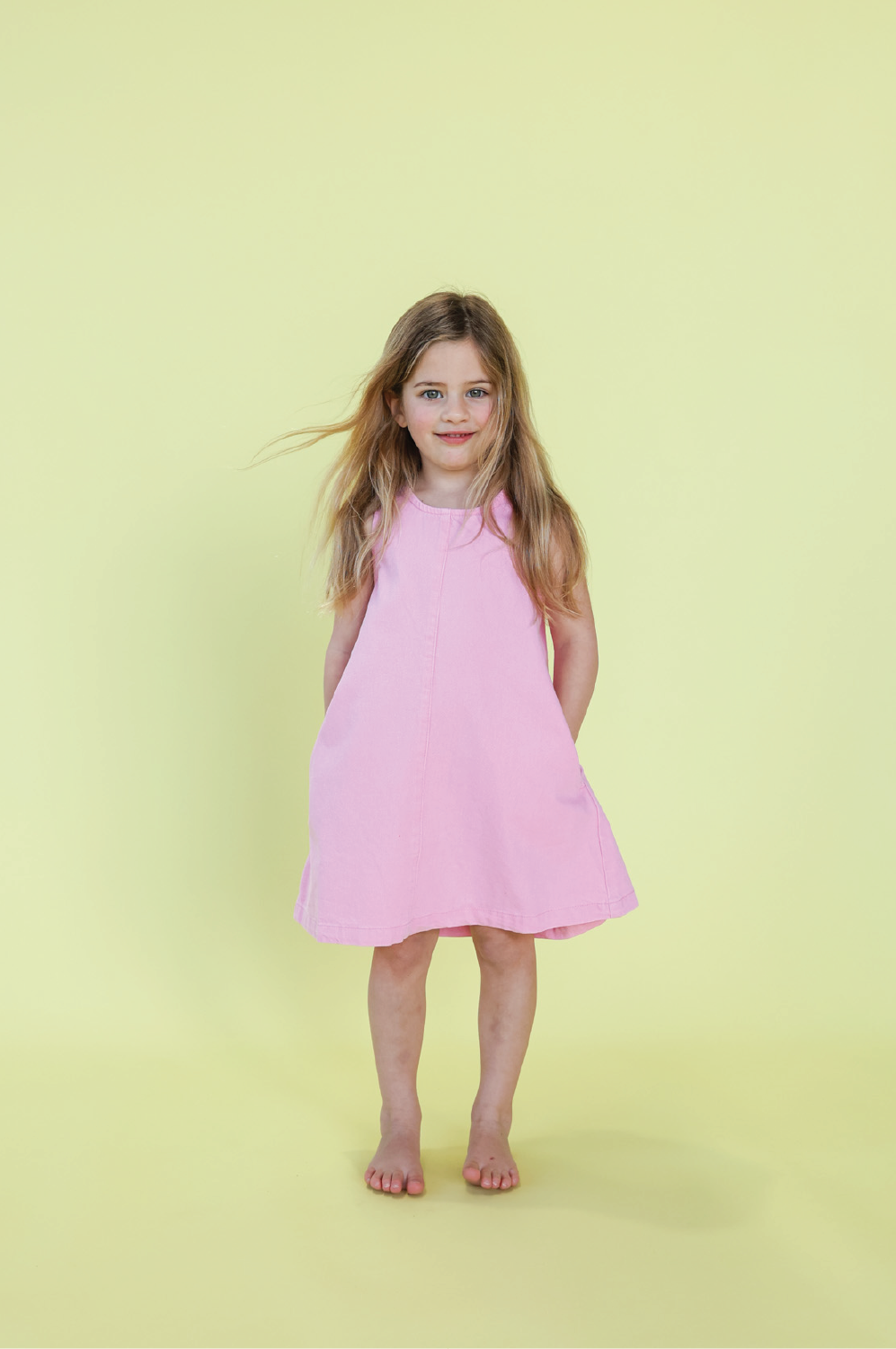 A young girl with long light brown hair stands barefoot against a pale yellow background, smiling slightly at the camera, wearing the GROWN - Organic Shift Denim Dress Candy by GROWN.