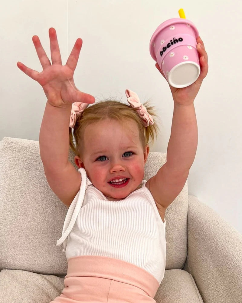 A smiling toddler with blonde pigtails sits on a light chair, wearing a white top and pink pants, raising one hand while holding a BBCINO BBcino Reusable Babycino Cup 120ml Daisy Baby Pink in the other.