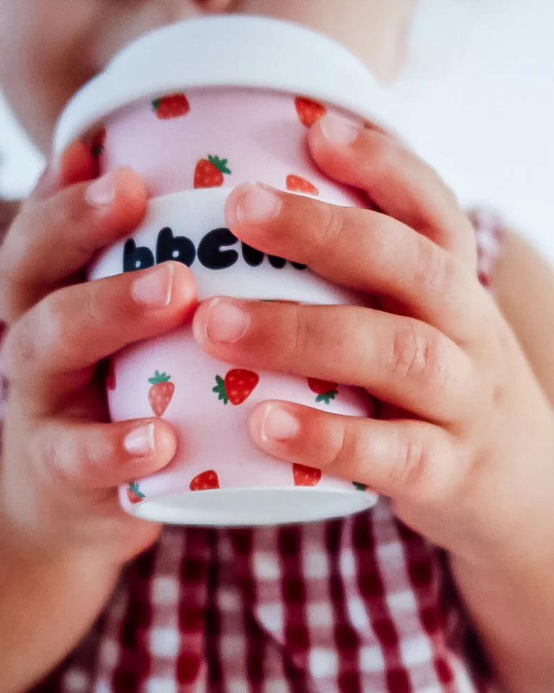 A child holds the BBCINO BBcino Reusable Babycino Cup 120ml Berry BB—an eco-friendly pink cup with a strawberry pattern and white lid—in close-up while wearing a red and white checkered dress.
