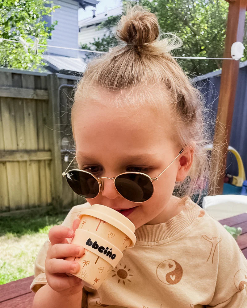 A young child with blonde hair in a top knot wears round sunglasses and a beige shirt, smiling while holding the BBCINO BBcino - Reusable Babycino Cup 120ml Oasis outdoors near a wooden fence.