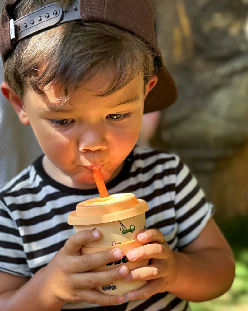 A young child wearing a backward brown cap and striped shirt drinks outdoors from the BBCINO BBcino - Reusable Babycino Cup 120ml Truck That, holding the eco-friendly cup with an orange straw in both hands.