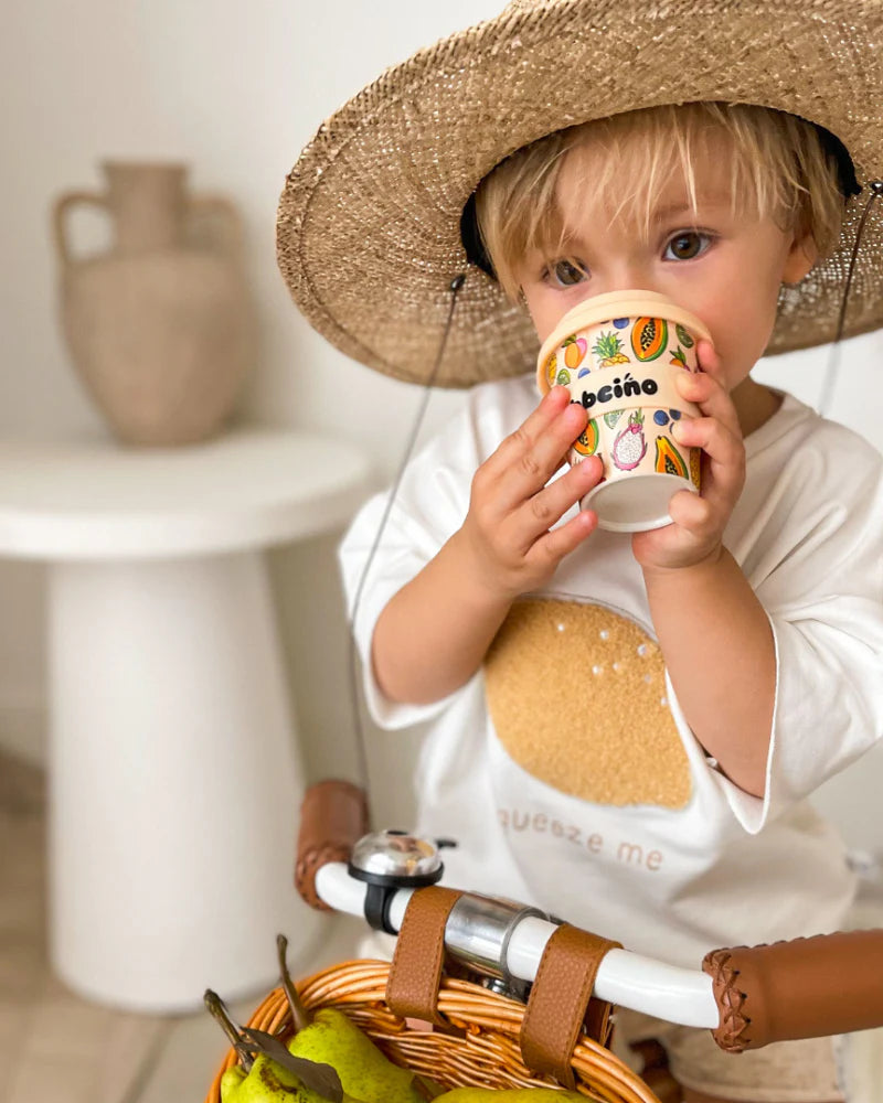A young child in a straw hat sips from the BBCINO BBcino - Reusable Babycino Cup 120ml Troppo (Limited Edition), decorated with fruit designs, while seated on a bicycle with pears in its basket, with a ceramic vase on a round white table behind.
