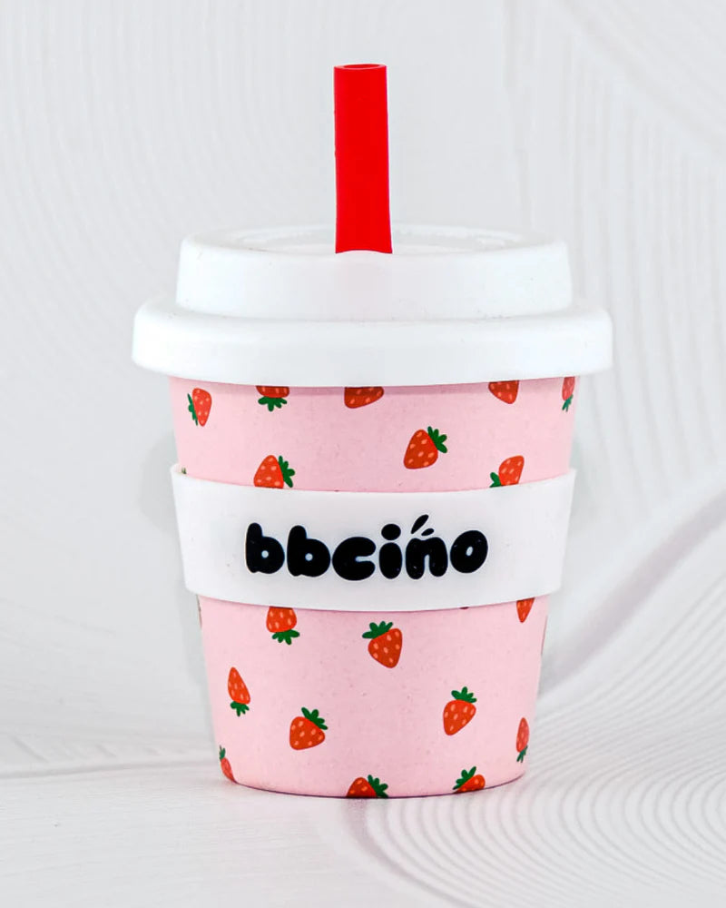 The BBcino - Reusable Babycino Cup 120ml Berry BB by BBCINO is a pink eco-friendly cup with a white lid, red straw, strawberry prints, and a white sleeve featuring "bbcino" in black lettering.