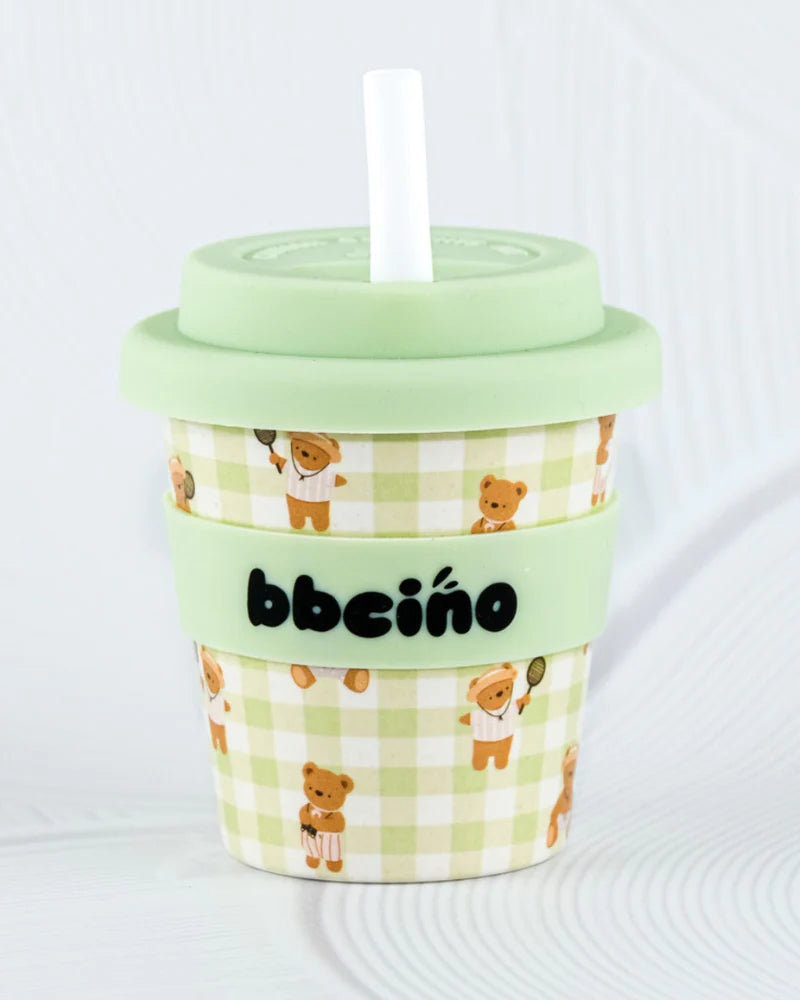 The BBcino - Reusable Babycino Cup 120ml Cub by BBCINO features a green lid, white straw, a green and white checkered pattern with teddy bears holding tennis rackets, and "bbcino" on a silicone band. Eco-friendly and reusable.