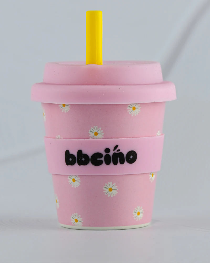 The BBCINO BBcino Reusable Babycino Cup 120ml Daisy Baby Pink features eco-friendly pink design with white daisy patterns, a yellow straw, pink lid, and a pink sleeve printed with "bbciño" in black text.