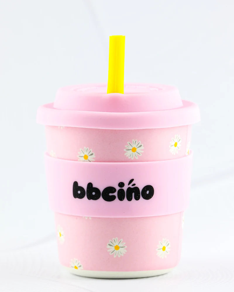 The BBCINO BBcino Reusable BambinoCino Cup 240ml Daisy Baby features a pink bamboo cup with white daisies, yellow straw, pink lid, and a pink band printed with "bbcino" in black. Made from sustainably farmed bamboo.
