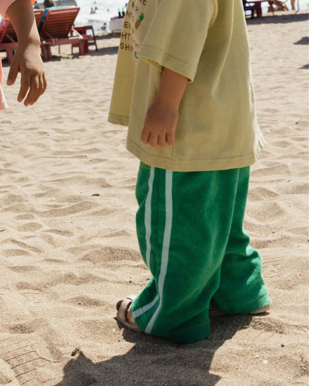 A young child in a yellow shirt and LENN LABEL's Weekend Terry Pant Jungle stands barefoot on the sandy beach, while another person’s hand reaches in from the left. Red lounge chairs and people appear in the background.