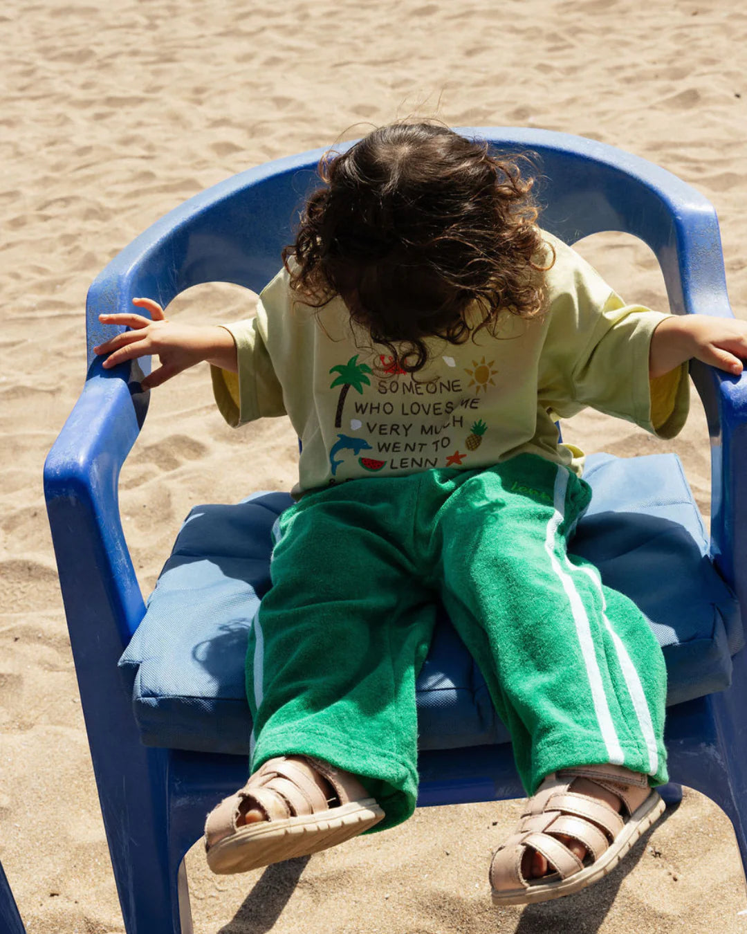 A young child with curly hair sits on a blue plastic chair at the beach, wearing a yellow t-shirt and the LENN LABEL Lenn Label - Weekend Terry Pant Jungle. The relaxed fit green pants complete their outfit as sand covers the ground in the background.