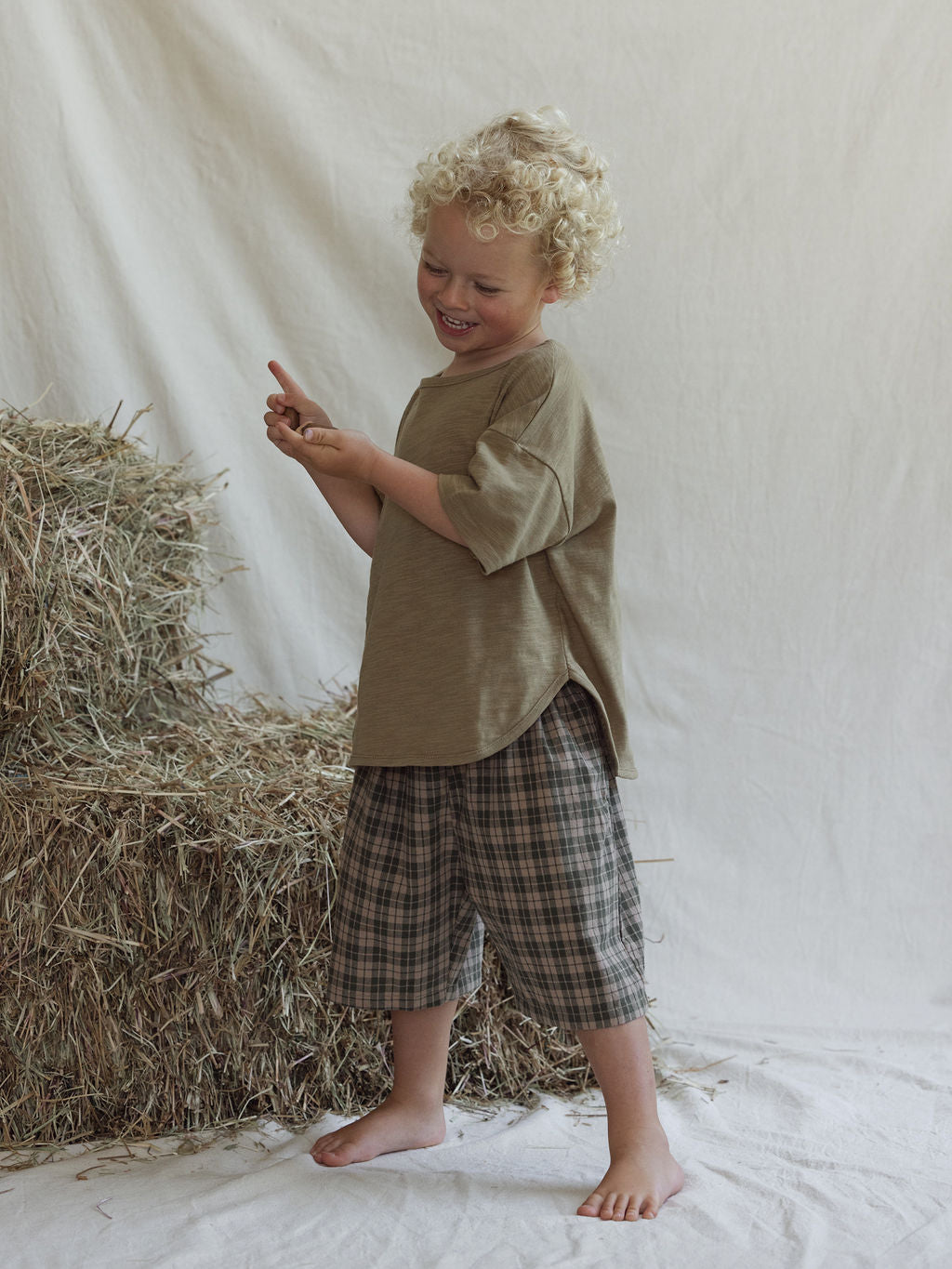 A young child with curly blond hair, wearing an oversized light brown unisex cotton shirt and VALENCIA BYRON BAY Farm Short Myrtle (Size 6), stands barefoot on a white cloth by hay bales, smiling and holding up one finger.