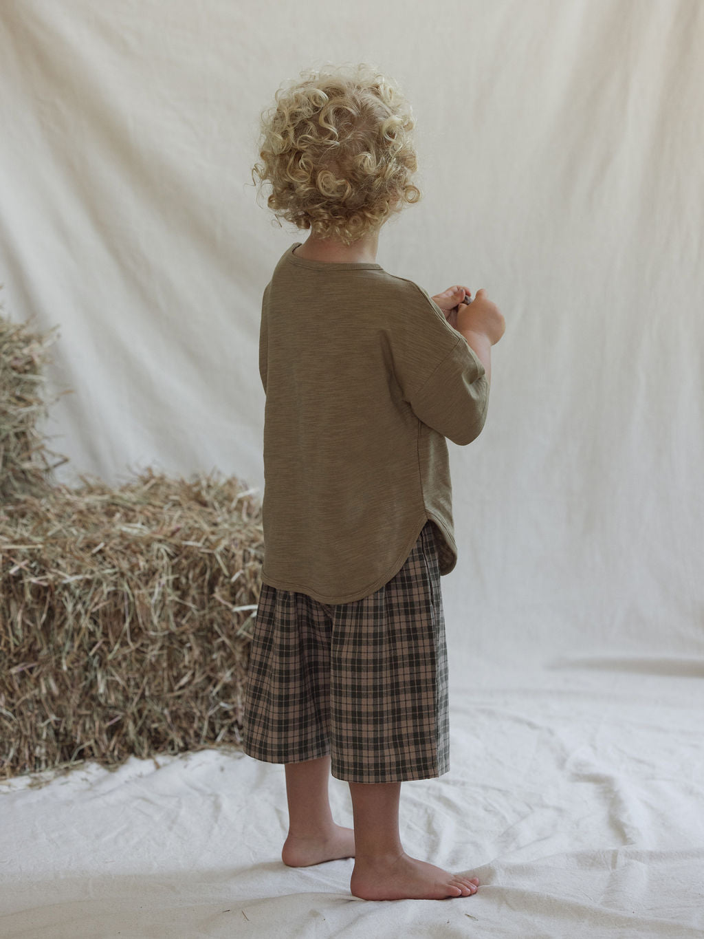 A young child with curly blond hair, barefoot on a white sheet and facing away, wears a loose beige top and VALENCIA BYRON BAY Farm Short Myrtle (Size 6), standing before stacked hay against a light fabric backdrop.