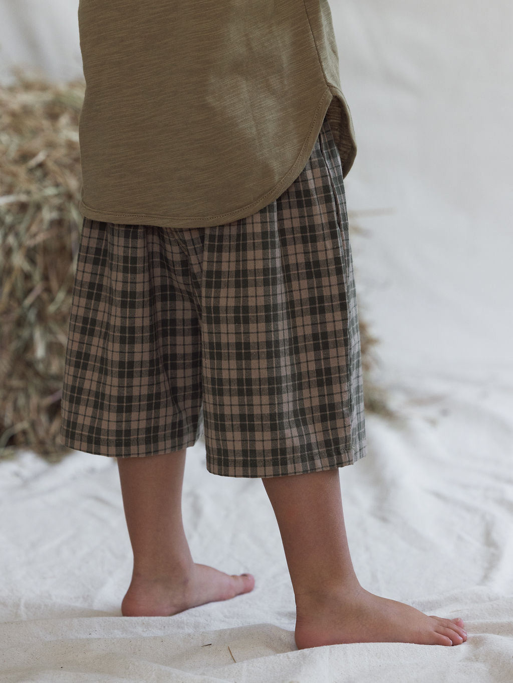 A child in a green unisex shirt from the VALENCIA BYRON BAY collection and plaid shorts stands barefoot on a light fabric surface, with a stack of hay visible in the background. The earthy tone of the Farm Short Myrtle shirt enhances the natural setting beautifully.