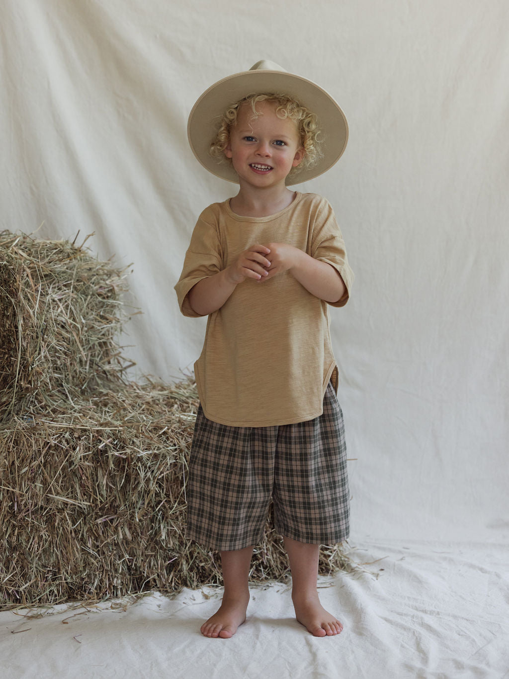 A young child with curly hair stands barefoot on a white cloth in front of a stack of hay, wearing the Farm Short Myrtle from VALENCIA BYRON BAY's unisex collection. Their wide-brimmed hat adds to the charm as they smile at the camera, framed by a simple white fabric backdrop.