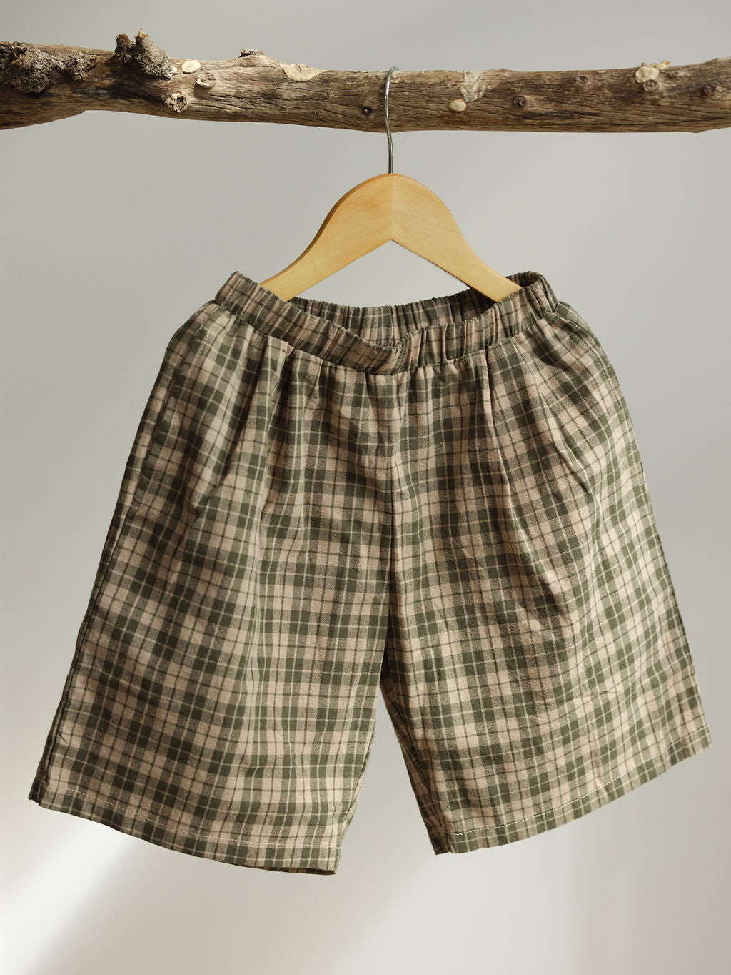 Farm Short Myrtle by VALENCIA BYRON BAY, crafted with durable cotton and designed in an earthy green tone and brown checkered pattern, hang on a wooden hanger supported by a natural branch against a neutral background. These plaid shorts, featuring an elastic waistband, are a standout piece in the unisex collection.