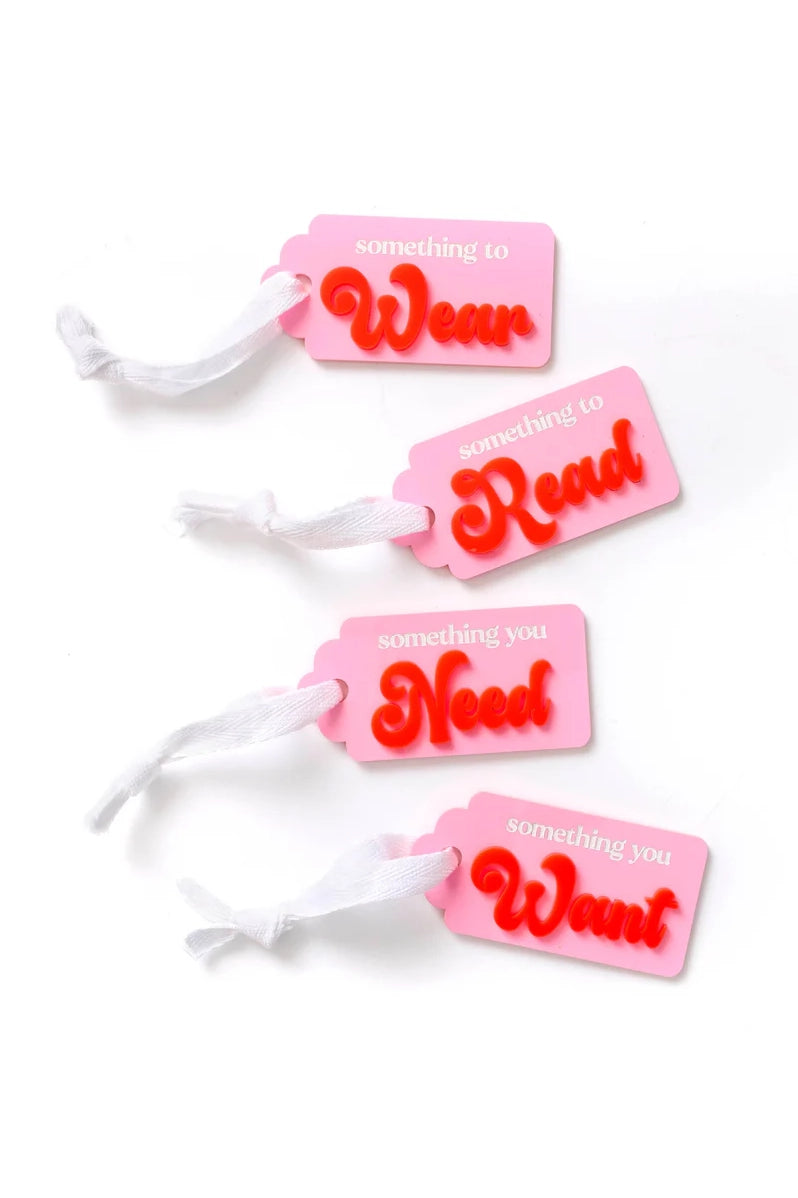 Four acrylic gift tags from HELLO KIT CO, named "WEAR, WANT, NEED, READ Tags Pink/Red," are arranged vertically with white ribbons. Each pink tag beautifully displays the phrases "something to Wear," "something to Read," "something you Need," and "something you Want" in striking red and white letters, providing a charming nod to traditional gifting practices.