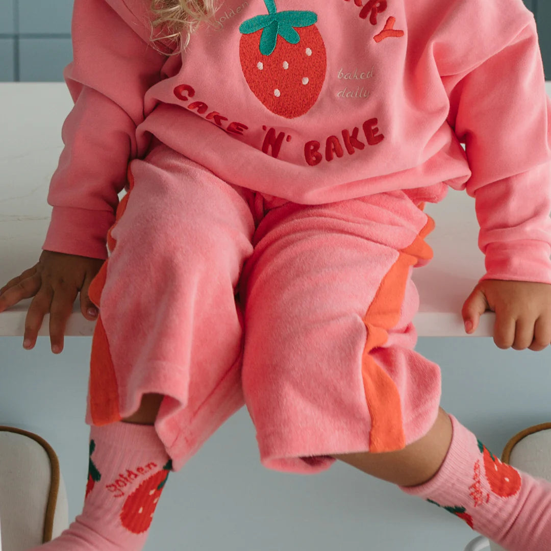 A child wears GOLDEN CHILDREN's Strawberry Cake Wide Leg Pants Terry Towel in Meringue Pink/Napoli, paired with a strawberry "Cake N' Bake" top and pink strawberry socks, sitting on a white surface.