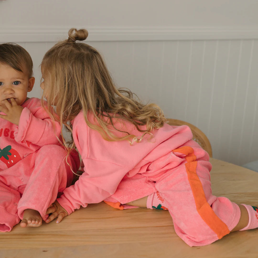 Two young children, dressed in GOLDEN CHILDREN's Strawberry Cake Wide Leg Pants in Meringue Pink/Napoli terry towel cotton, sit on a wooden table—one whispering to the other, who smiles at the camera with fingers to their mouth.