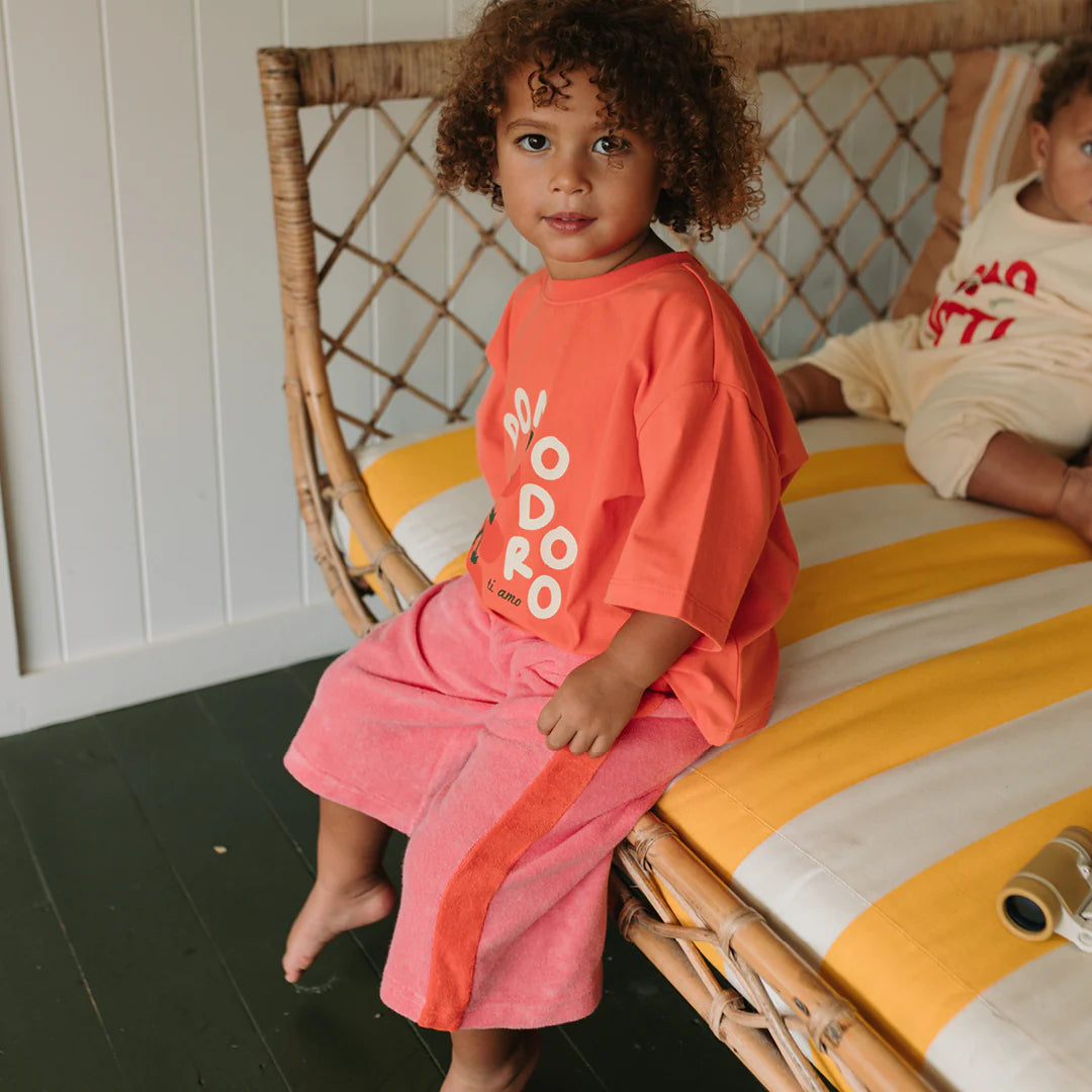 A young child with curly hair sits on a yellow and white striped cushioned wicker bench, wearing a coral t-shirt and GOLDEN CHILDREN - Strawberry Cake Wide Leg Pants Terry Towel Meringue Pink/Napoli. Another child is partially visible beside them.