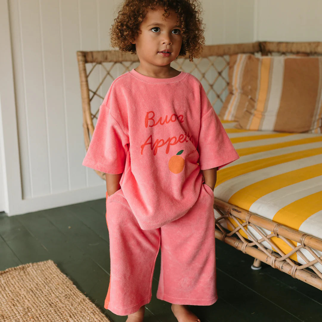 A child with curly hair wears GOLDEN CHILDREN's Strawberry Cake Wide Leg Pants Terry Towel in Meringue Pink/Napoli, standing indoors by a rattan sofa with yellow and white striped cushions.
