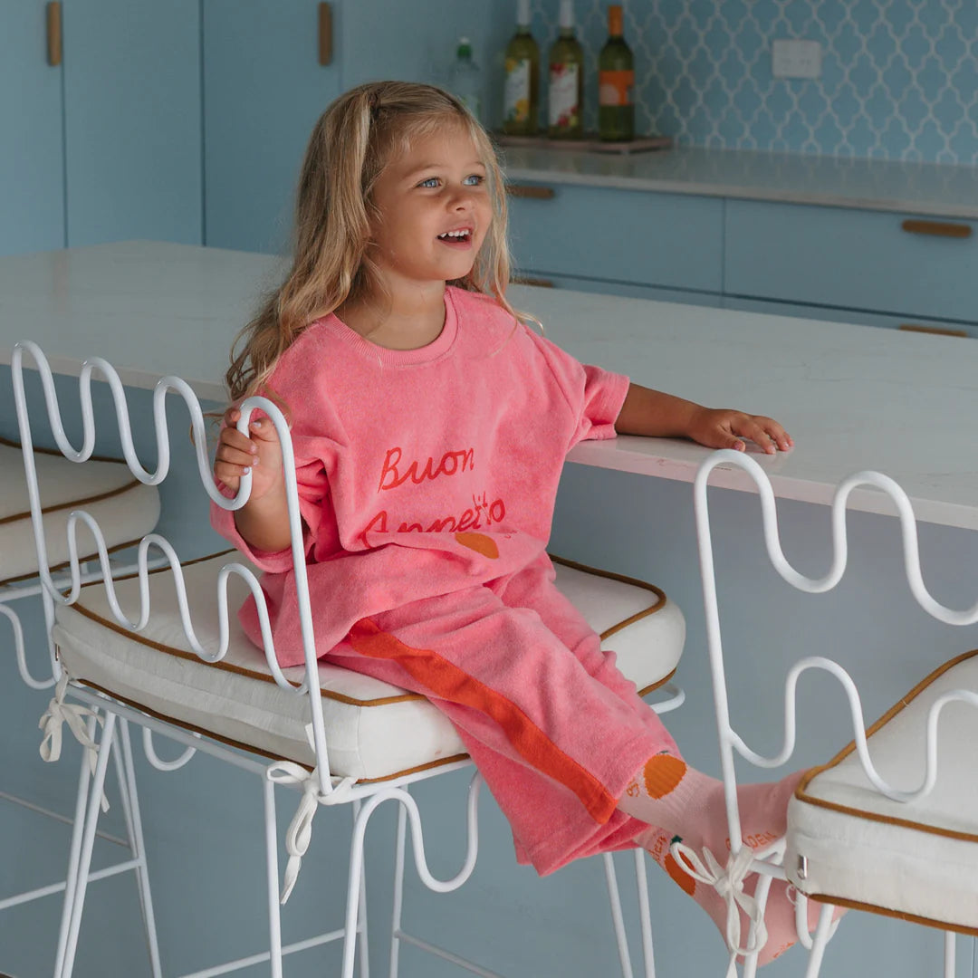 A young child with long hair sits sideways on a white barstool in a light blue kitchen, smiling and wearing GOLDEN CHILDREN's Strawberry Cake Wide Leg Pants Terry Towel Meringue Pink/Napoli, paired with a pink “Buon Appetito” shirt.