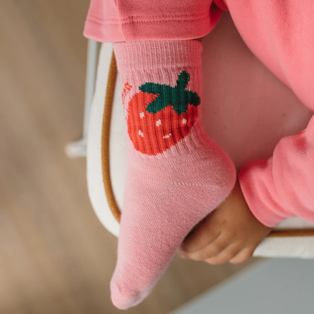 A child in pink pajamas wears GOLDEN CHILDREN's Golden Children - Strawberry Cake Socks Strawberry with a red strawberry design, sitting on a white chair and resting their foot on the seat.
