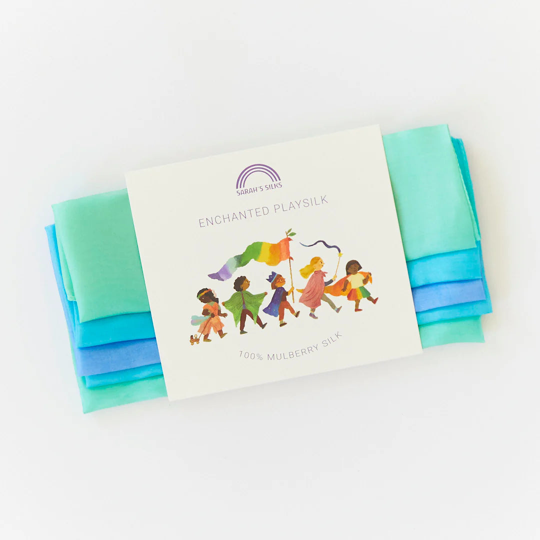 The "Enchanted Playsilk Sea" by SARAH'S SILKS is crafted from 100% mulberry silk in captivating shades of blue and green, designed to ignite creativity. Its packaging includes an illustration of children exploring their imagination with the colorful fabric.