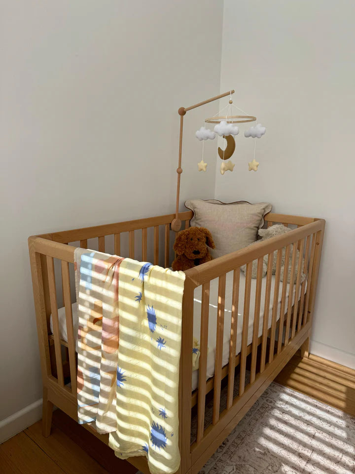 A wooden baby crib with pillows, a teddy bear, and ROOMI & ME’s Baby Mobile Starry Night for visual stimulation. Sunlight casts striped shadows across the crib and a yellow blanket with blue prints drapes over the side.