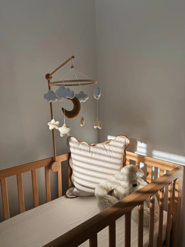 A wooden crib with the ROOMI & ME Baby Mobile Starry Night, featuring handmade clouds and moons, a cloud pillow, and a plush teddy bear. Sunlight casts soft shadows in the neutral-toned nursery for your little one's visual stimulation.