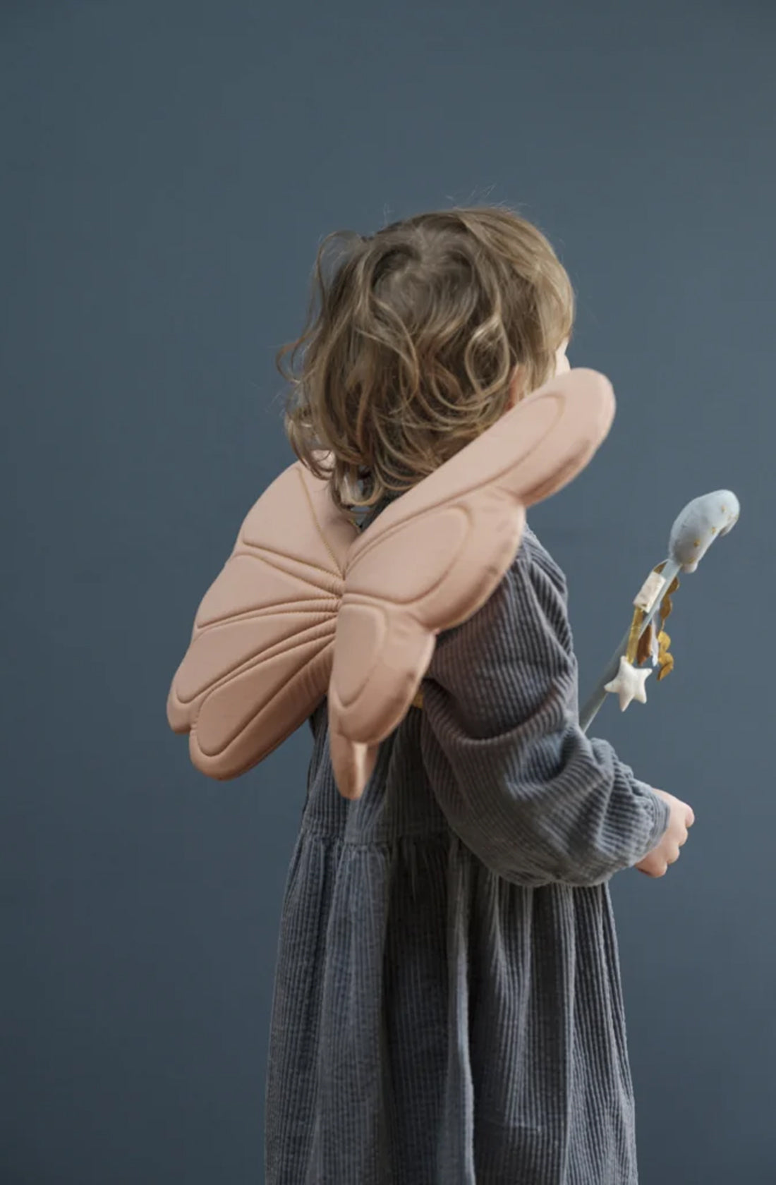 A child with wavy brown hair is seen from behind, wearing FABELAB's Dress Up Wings Old Rose and a dark grey long-sleeve dress. The child holds a stuffed grey bird with a wooden ring and a star attached, lost in the world of pretend play against a plain, dark grey wall.